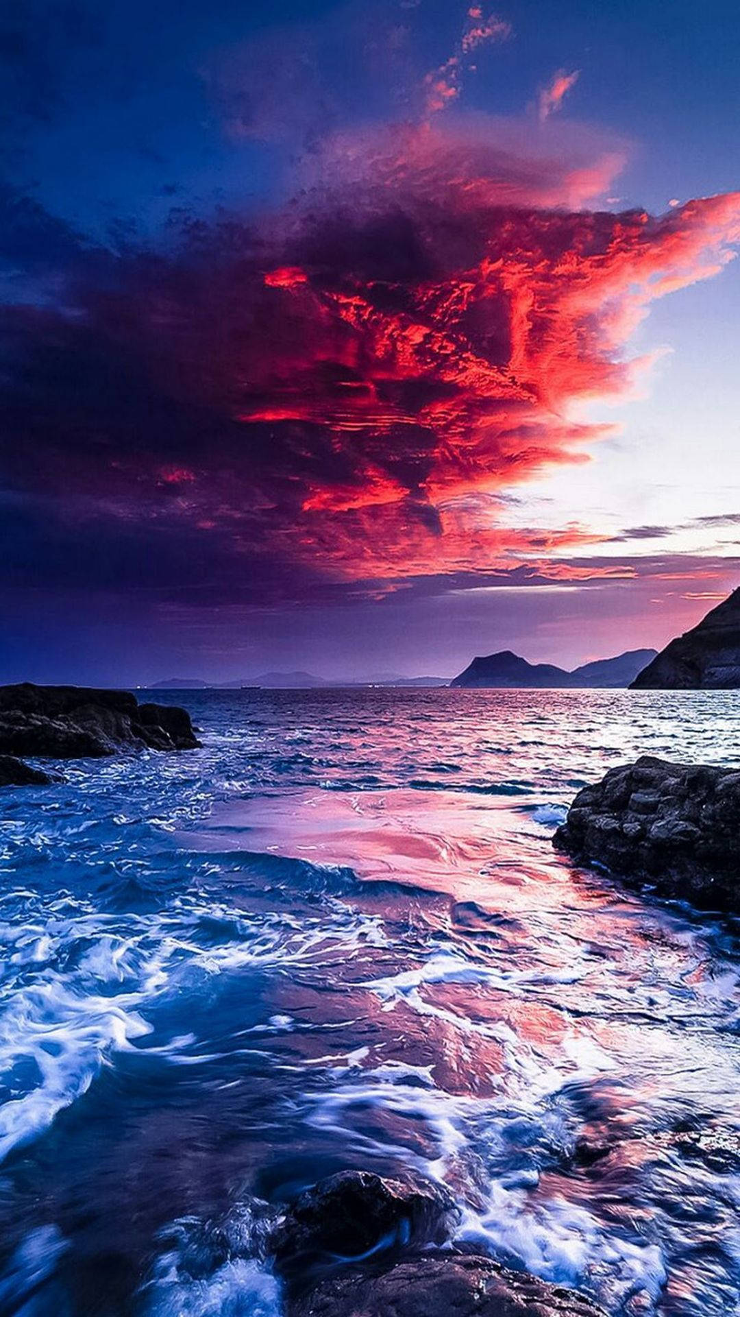 海灘 桌布 壁紙 — 鮮豔的紅色和紫色雲彩在岩石海岸線上燃燒，海浪在黃昏時拍打著暗色的石頭。