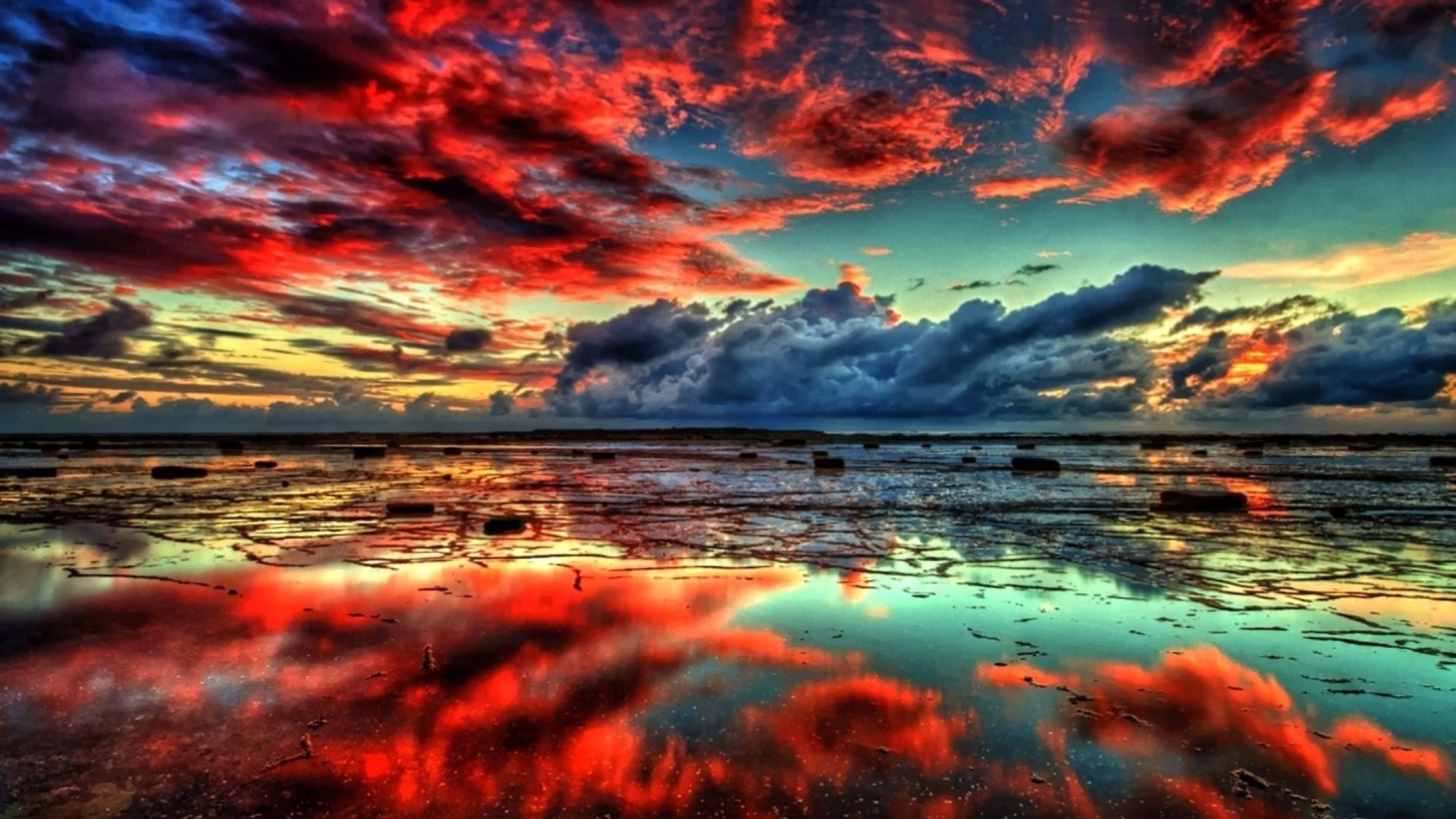 4K Display 桌布 壁紙 — 鮮豔的紅色和藍色雲彩在平坦地平線上的戲劇性日落中倒映在淺水中。