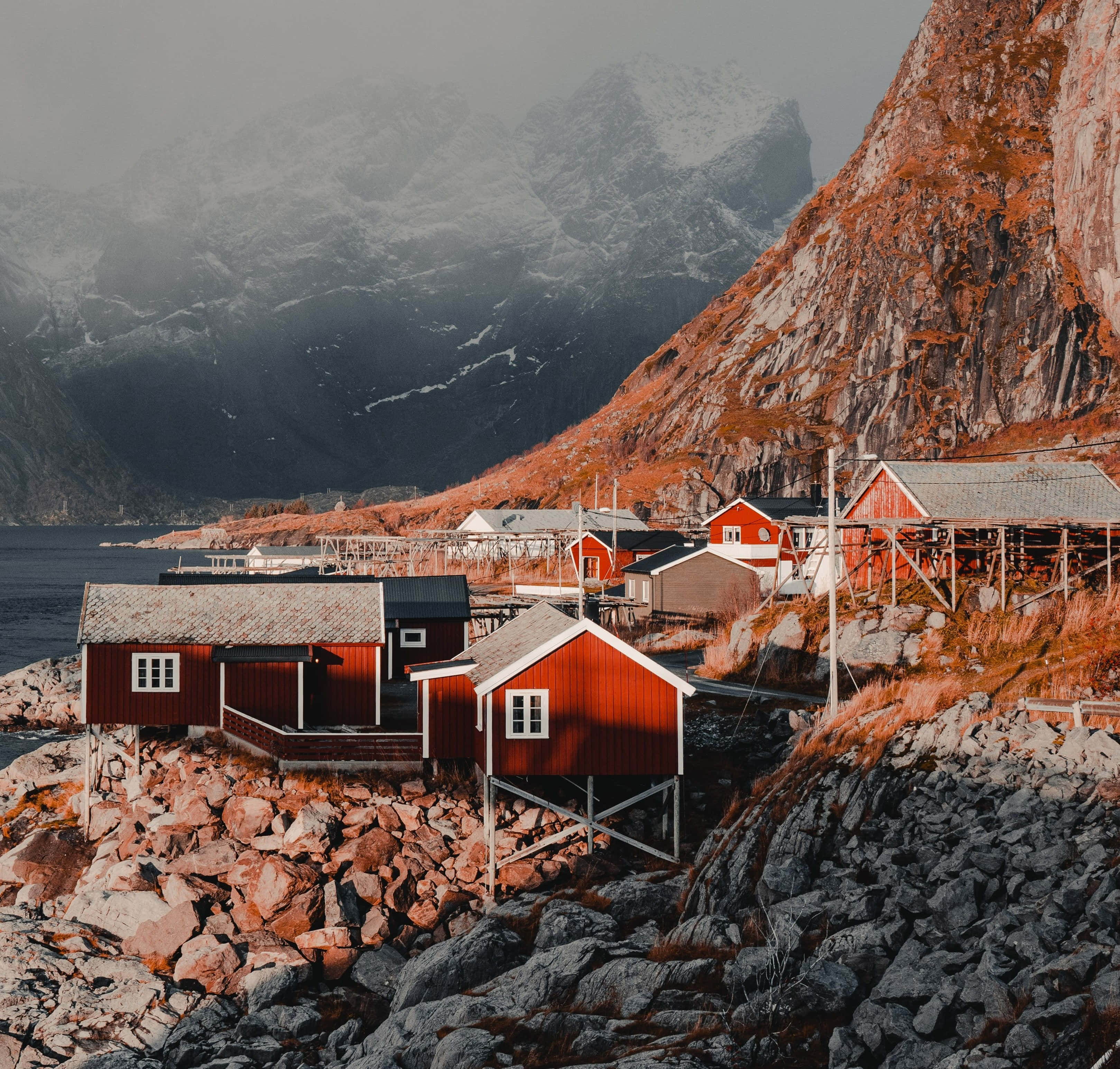 Norway 桌布 壁紙 — 鮮豔的紅色木屋坐落在峽灣旁的岩石懸崖上，白雪覆蓋的山脈在迷霧背景中若隱若現。
