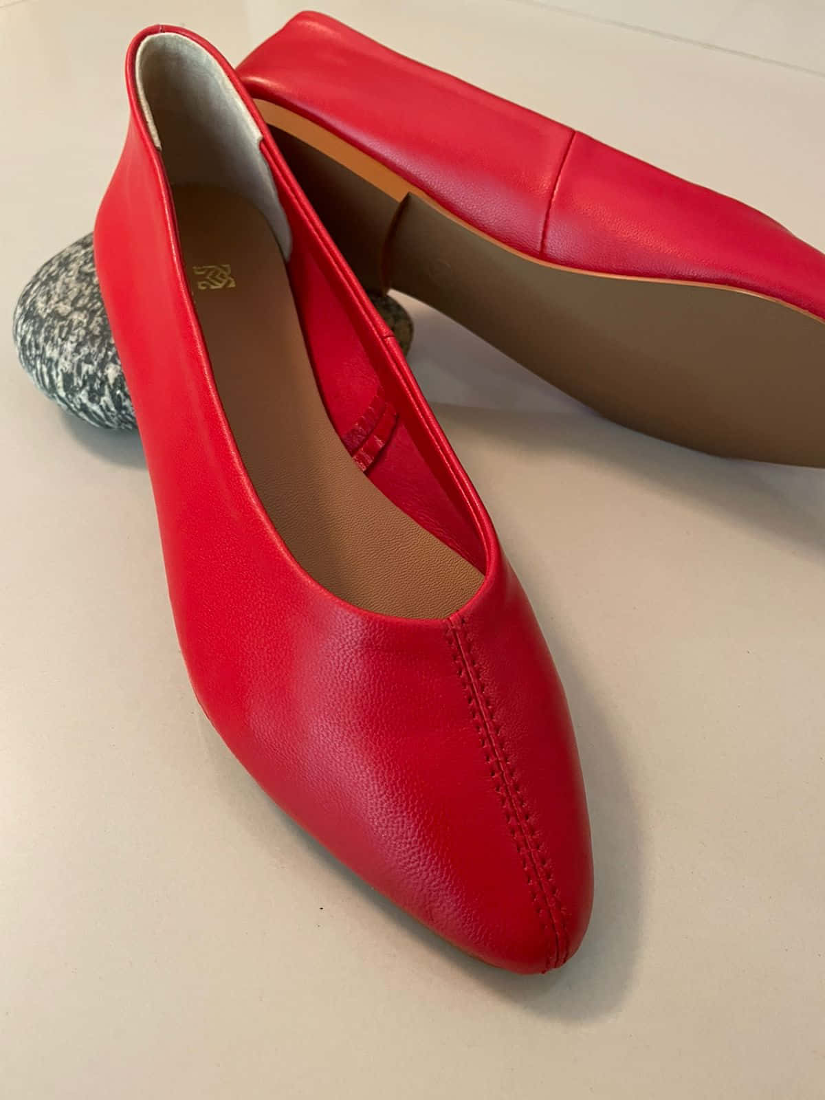 Red Shoes 桌布 壁紙 — 鮮豔的紅色皮革平底鞋，尖頭設計，放在淺色表面上，旁邊有一塊斑點石。