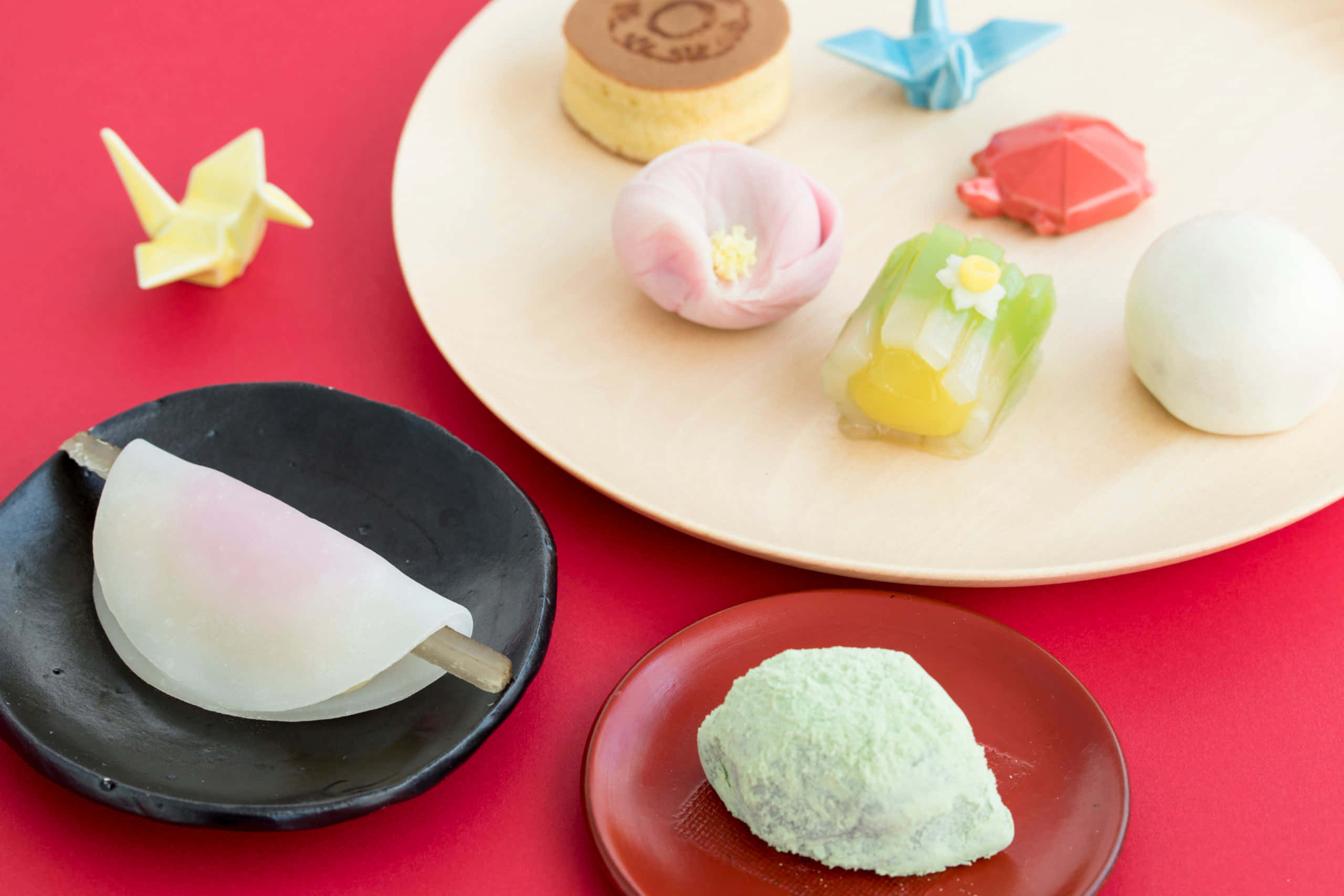 Japanese Sweets 桌布 壁紙 — 鮮豔的紅色表面上展示了木製和陶瓷盤子上的各種精緻日本甜點和摺紙鶴。