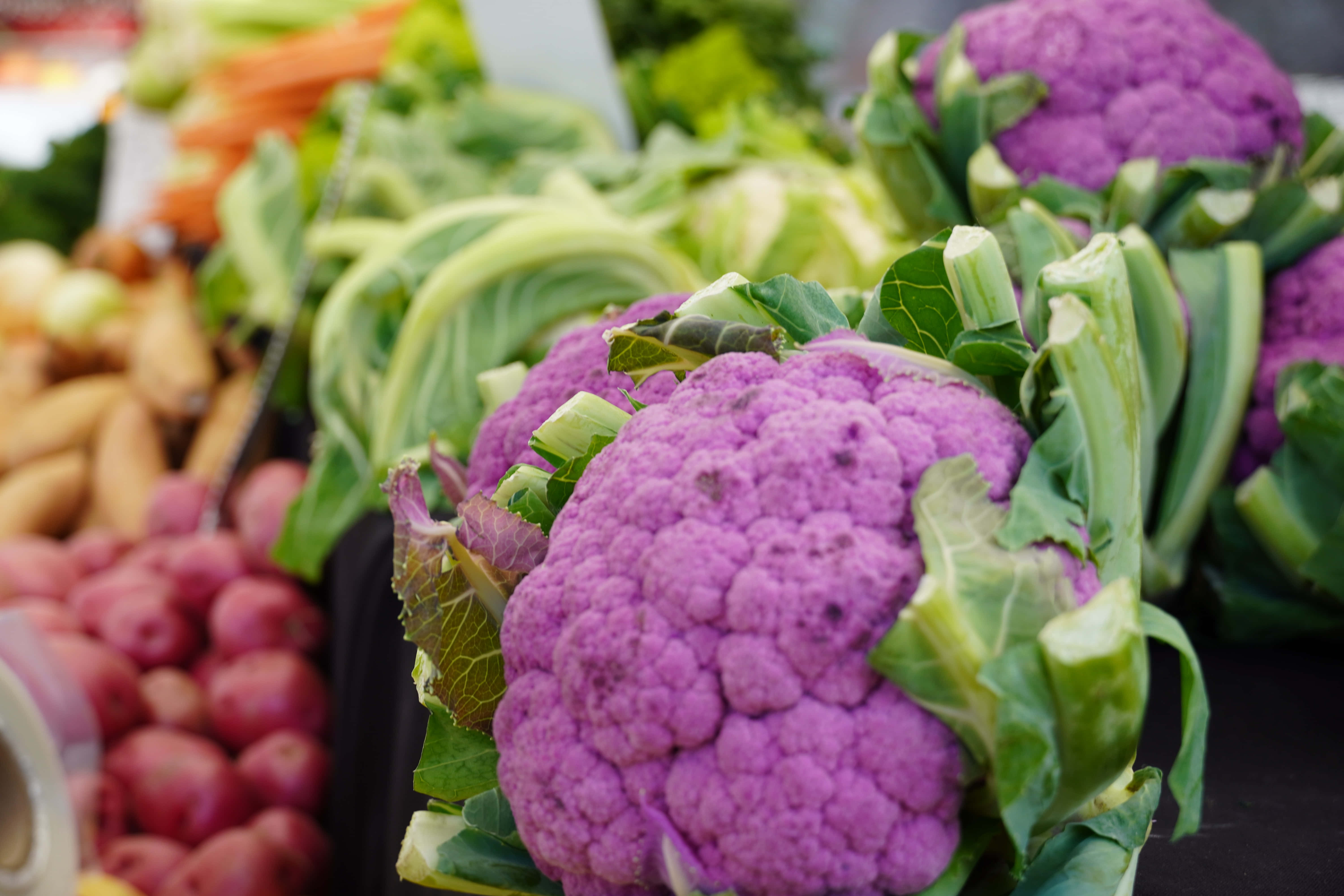 Purple Cauliflower 桌布 壁紙 — 鮮豔的紫色椰菜花配以綠色葉子，在市場攤檔的其他色彩繽紛的新鮮蔬菜中格外突出。