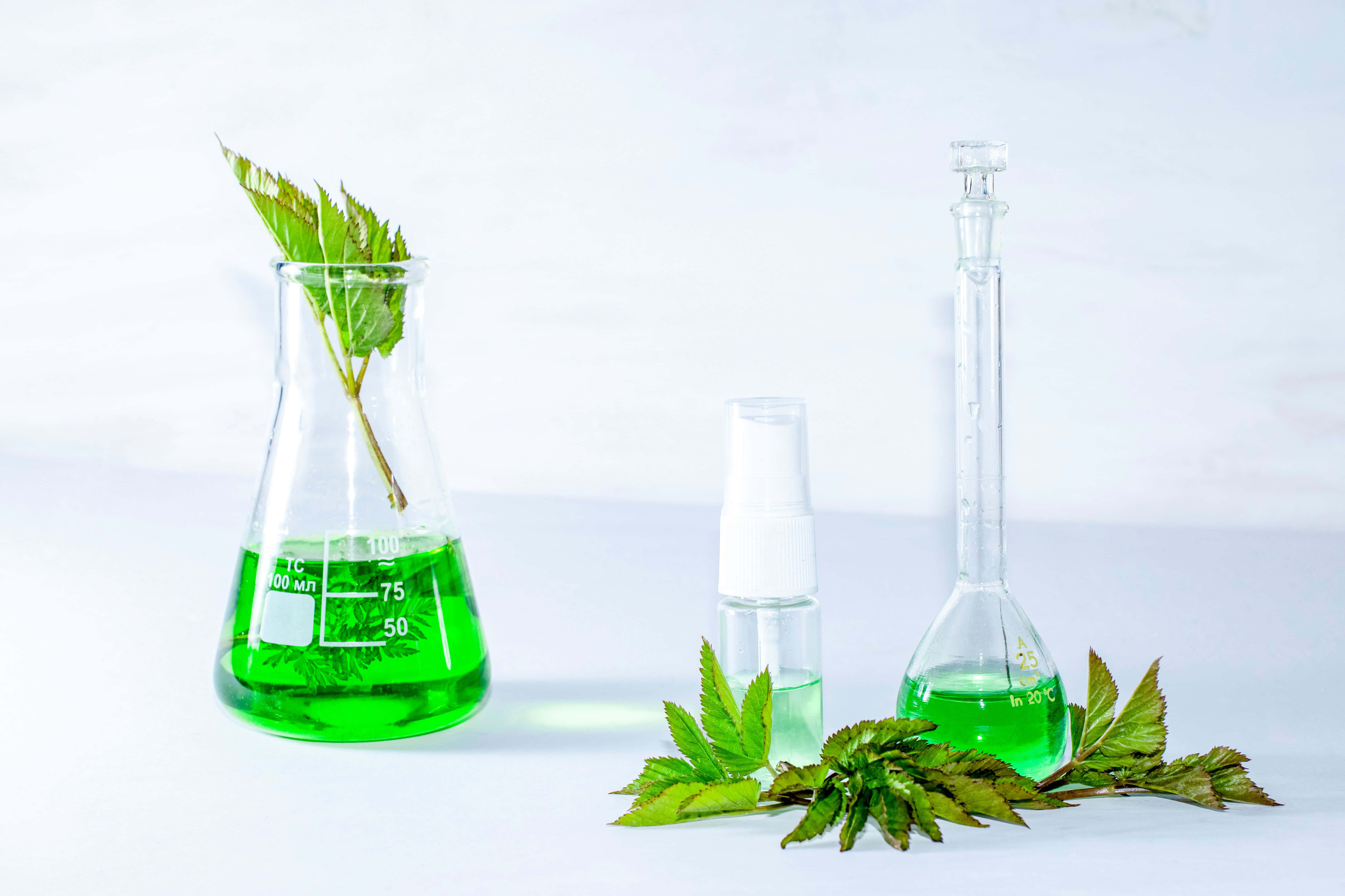Green Chemistry 桌布 壁紙 — 鮮豔的綠色液體裝滿玻璃實驗室燒瓶和一個噴霧瓶，旁邊是新鮮的綠葉，放在明亮的白色表面上。