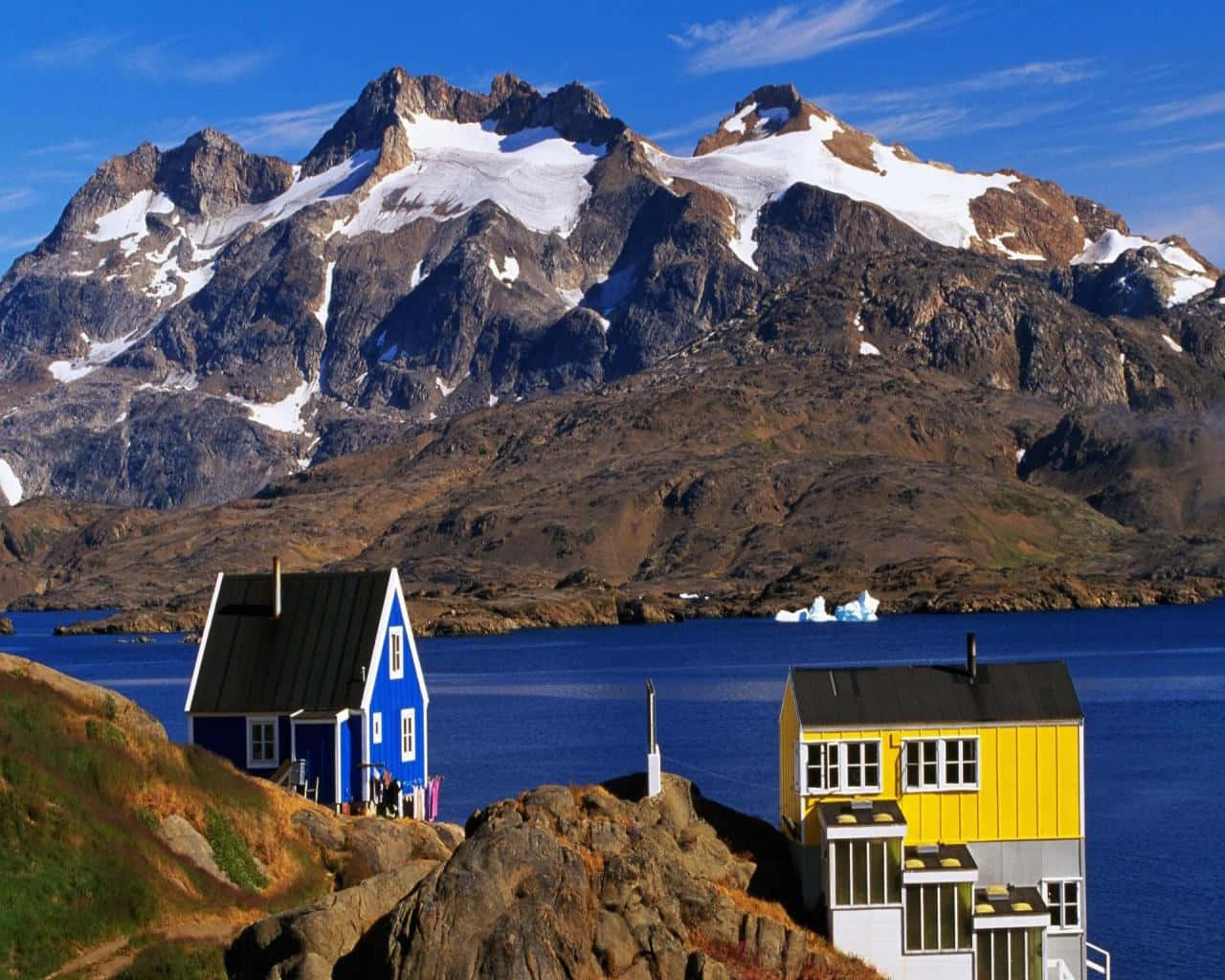 Greenland 桌布 壁紙 — 鮮豔的藍色和黃色房屋坐落在岩石懸崖上，俯瞰著深藍色的峽灣，遠處有白雪覆蓋的山脈和冰山。