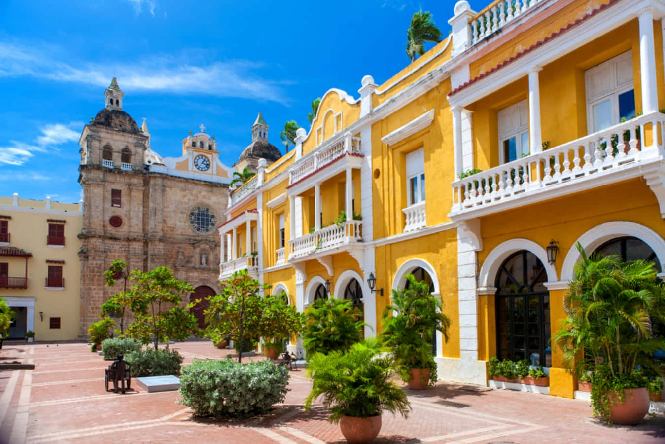 Colombia 桌布 壁紙 — 鮮豔的黃色殖民風格建築，帶有白色陽台和拱形門口，排列在陽光普照的廣場上，旁邊是一座歷史悠久的石頭教堂，在清澈的藍天下。