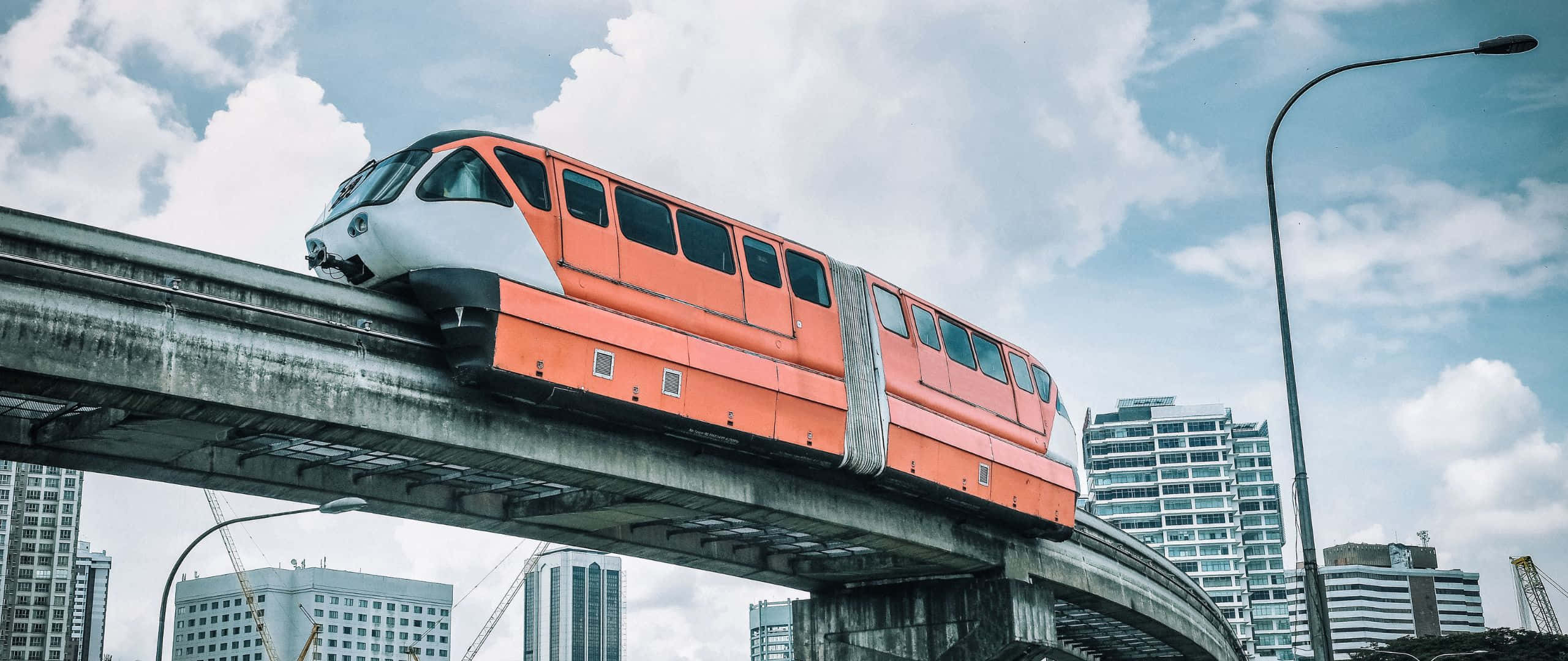 現代城市摩天大樓和多雲天空下，高架軌道上的橙色單軌列車。