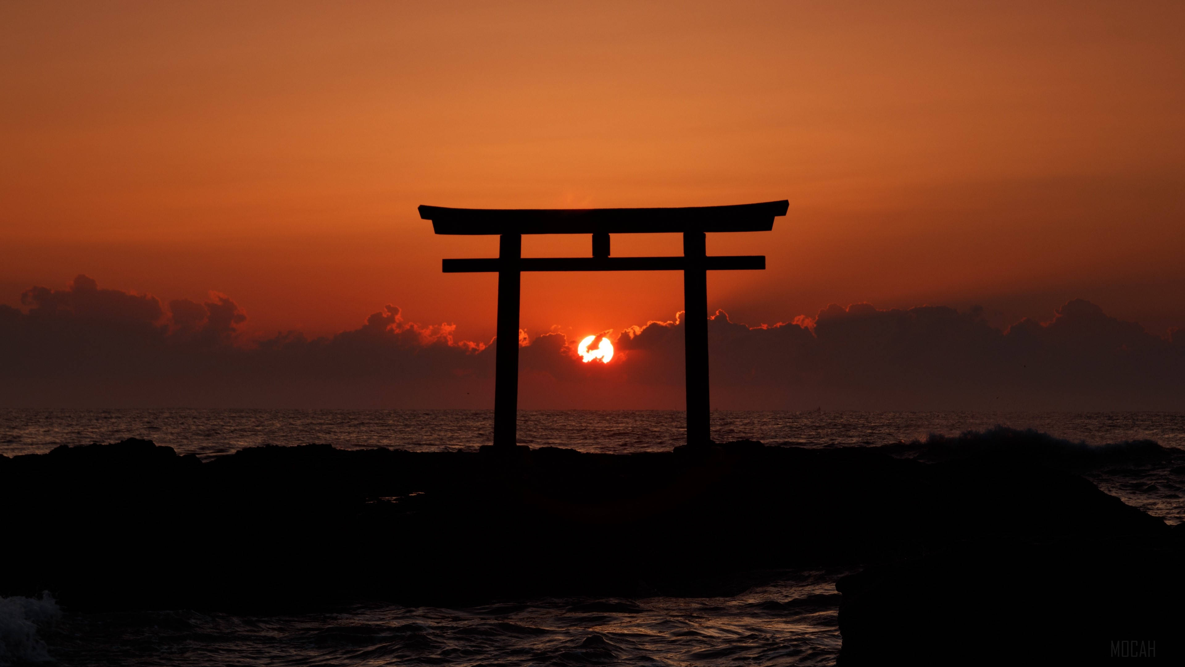 鳥居與夕陽 壁紙 — 一座剪影風格的日本鳥居矗立在岩石海岸上，夕陽在其後方以火橙色沉入海洋。