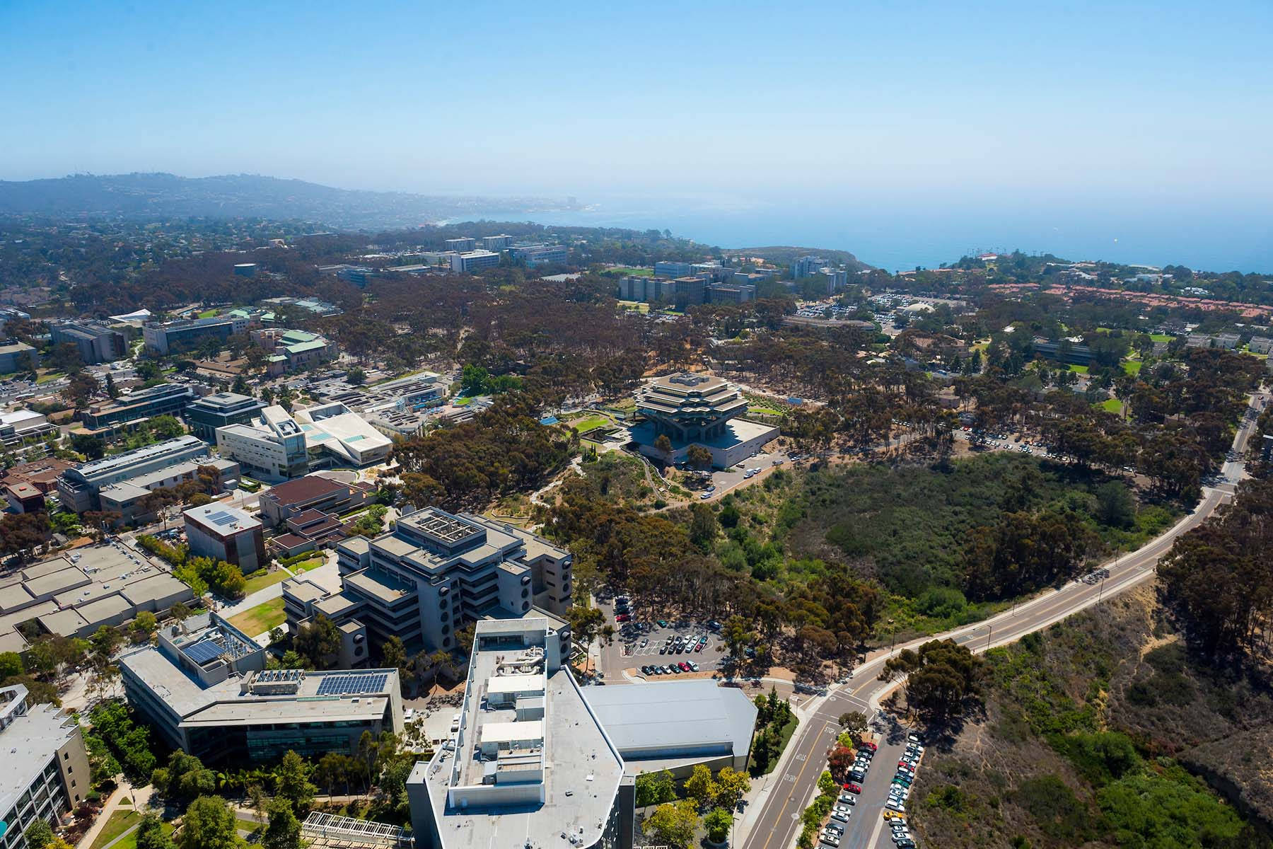 鳥瞰 UCSD 校園 壁紙 — 俯瞰廣闊嘅大學校園，被樹木環繞，遠方有海洋同山丘，天空晴朗蔚藍。