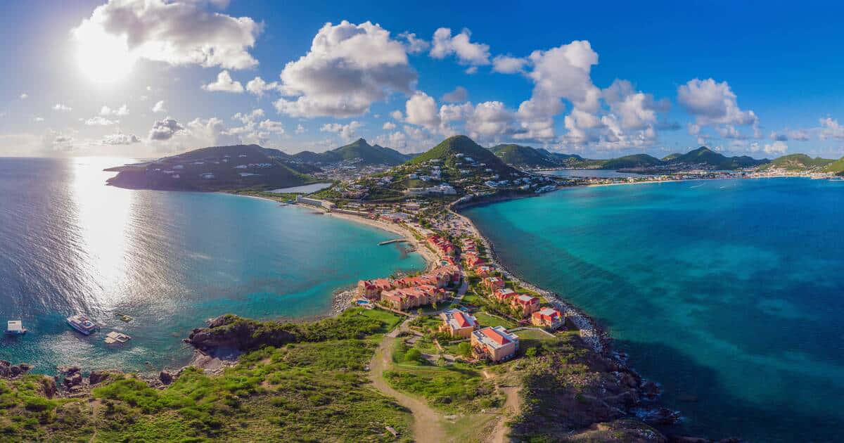 Caribbean Island 桌布 壁紙 — 鳥瞰一個充滿活力的沿海小鎮，坐落在碧綠的海水和鬱鬱蔥蔥的綠色山丘之間，明亮的藍天上有蓬鬆的雲朵。