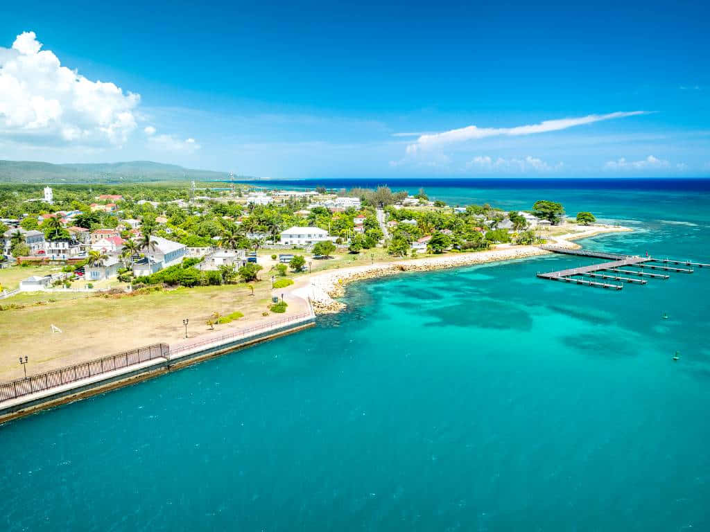 Jamaican Island 桌布 壁紙 — 鳥瞰一個沿海小鎮，擁有碧綠海水、碼頭和茂密綠植，上方是明亮的藍天。