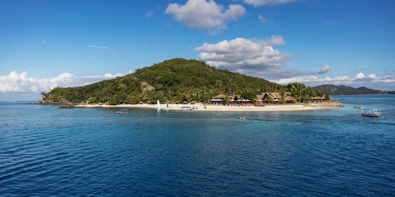 Caribbean Island 桌布 壁紙 — 鳥瞰一個鬱鬱蔥蔥的熱帶島嶼，擁有沙灘度假村建築和船隻，在明亮的天空下漂浮在充滿活力的藍色海洋中。