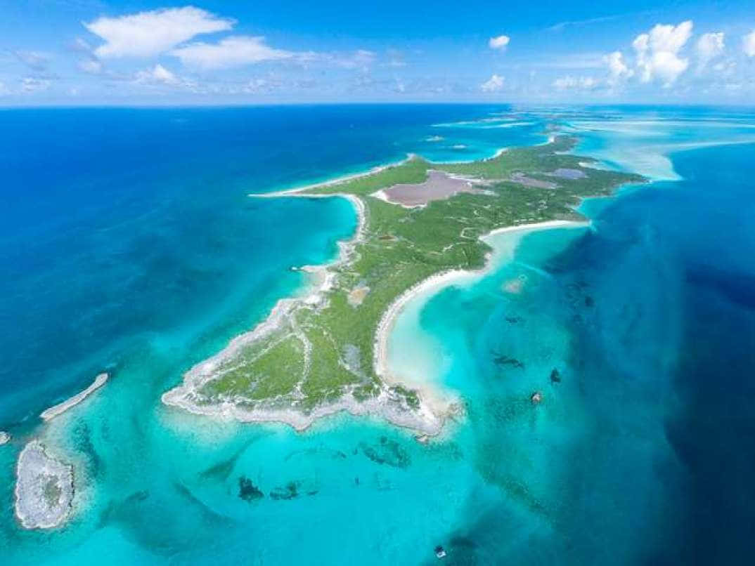 Bahamas Island 桌布 壁紙 — 鳥瞰一個鬱鬱蔥蔥的綠色島嶼，周圍環繞著充滿活力的碧綠海水，在明亮的藍天下有零星的白雲。