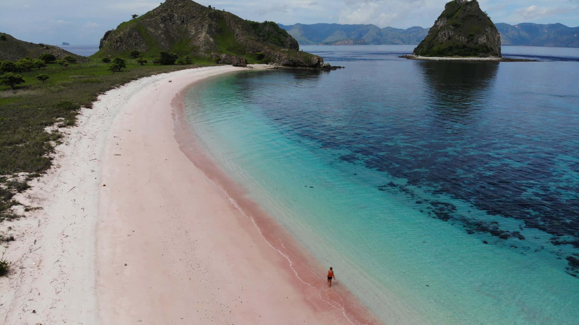 Pink Beach 桌布 壁紙 — 鳥瞰圖：一個人獨自走在迷人的粉紅沙灘上，遠處是碧綠的海水和岩石島嶼。