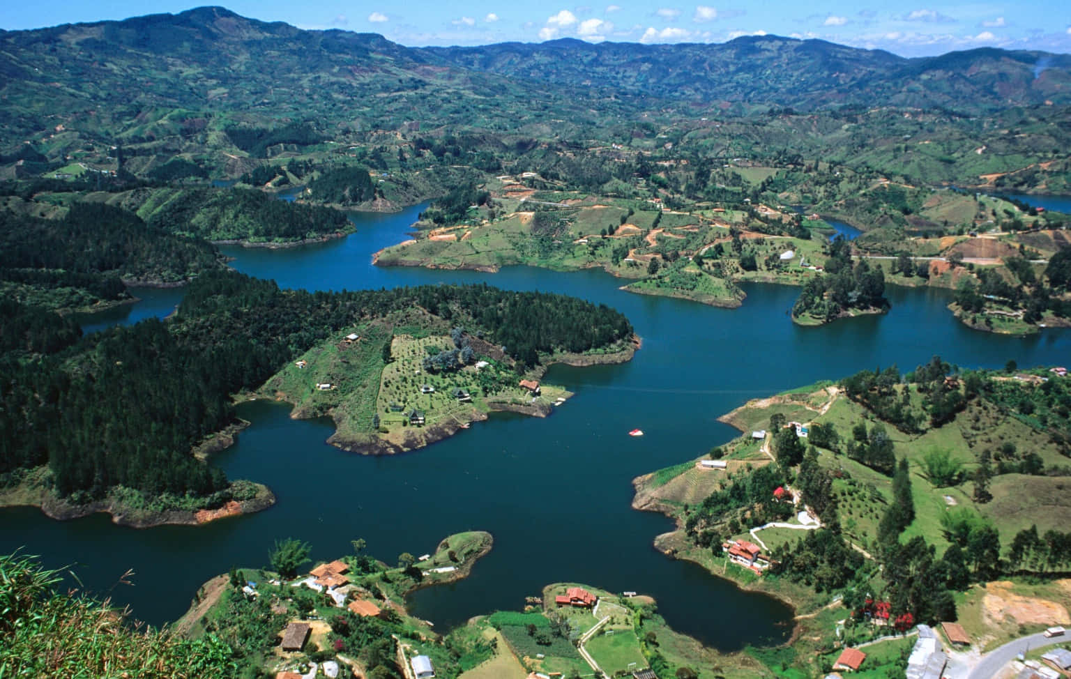 Colombia 桌布 壁紙 — 鳥瞰圖：在明亮的天空下，蜿蜒的藍色湖泊坐落在鬱鬱蔥蔥的綠色山丘之間，山丘上點綴著小房屋和樹木。