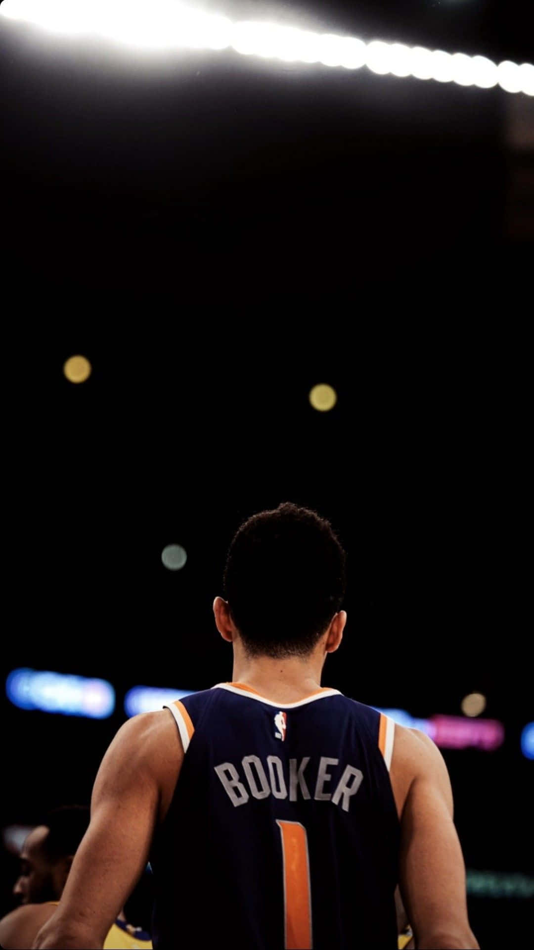 鳳凰城太陽隊球星德文·布克即將開戰 壁紙 — 一名身穿 'BOOKER 1' 球衣的籃球運動員在明亮的場館燈光下背對鏡頭站立。