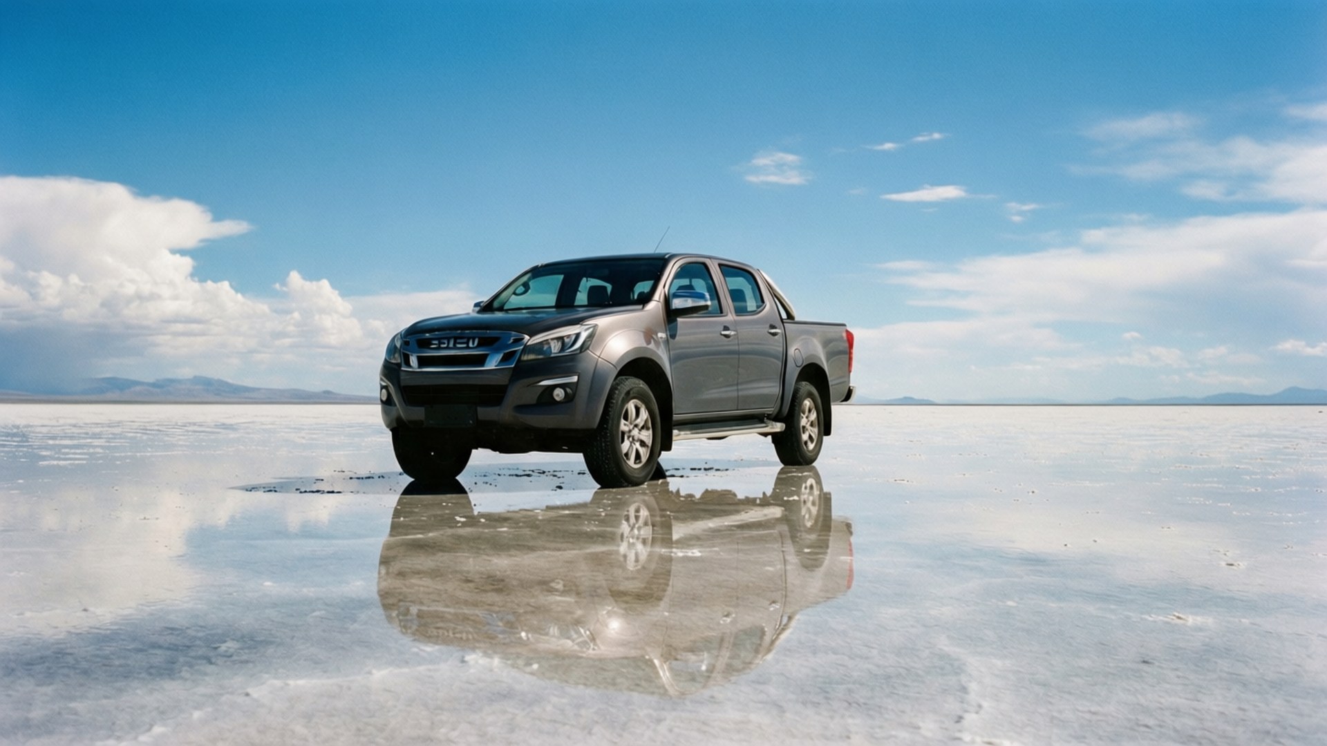鹽灘鏡像 壁紙 — 一架銀色五十鈴皮卡車站在廣闊的鹽灘上，在藍天白雲下反射其影像。