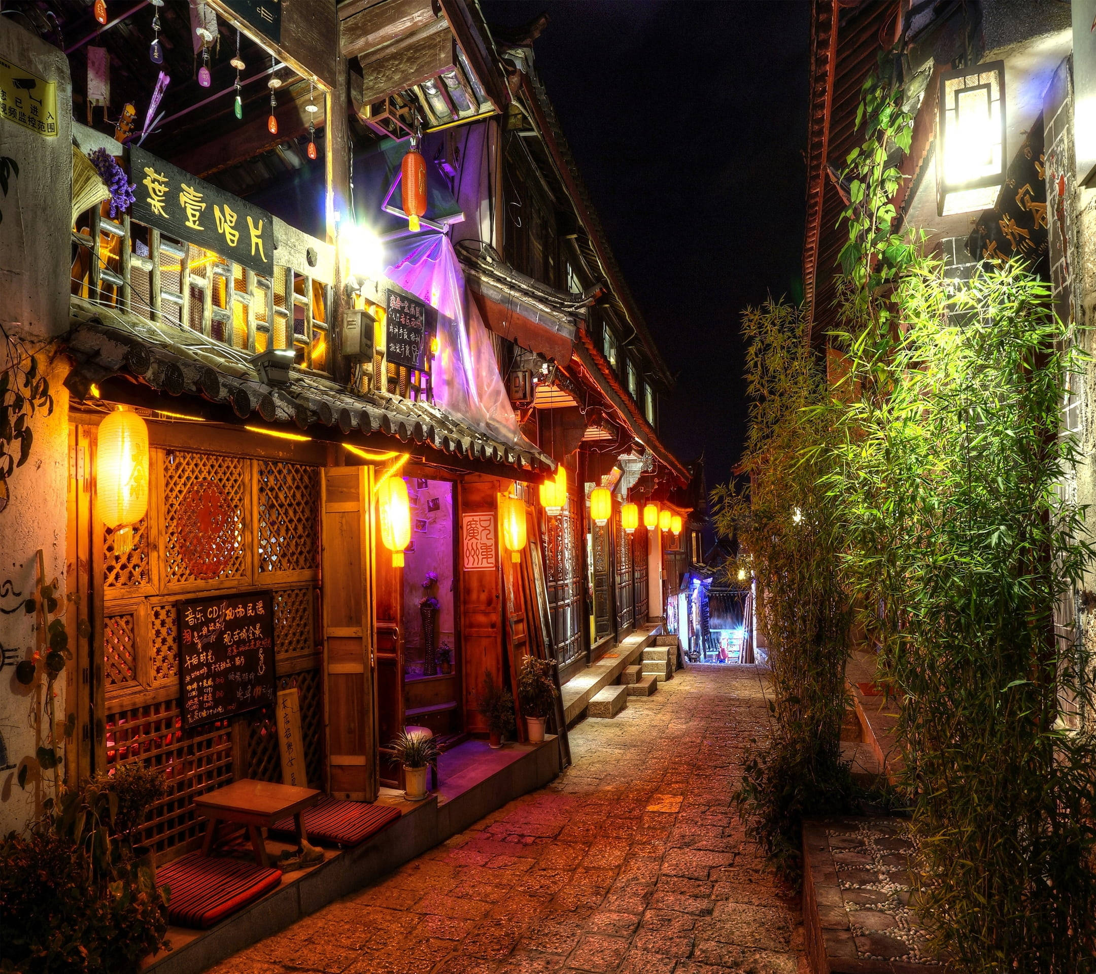 麗江古城夜景 壁紙 — 夜晚一條狹窄的鵝卵石小巷，被溫暖的燈籠和傳統木建築上發光的招牌照亮，建築上攀爬著綠色植物。
