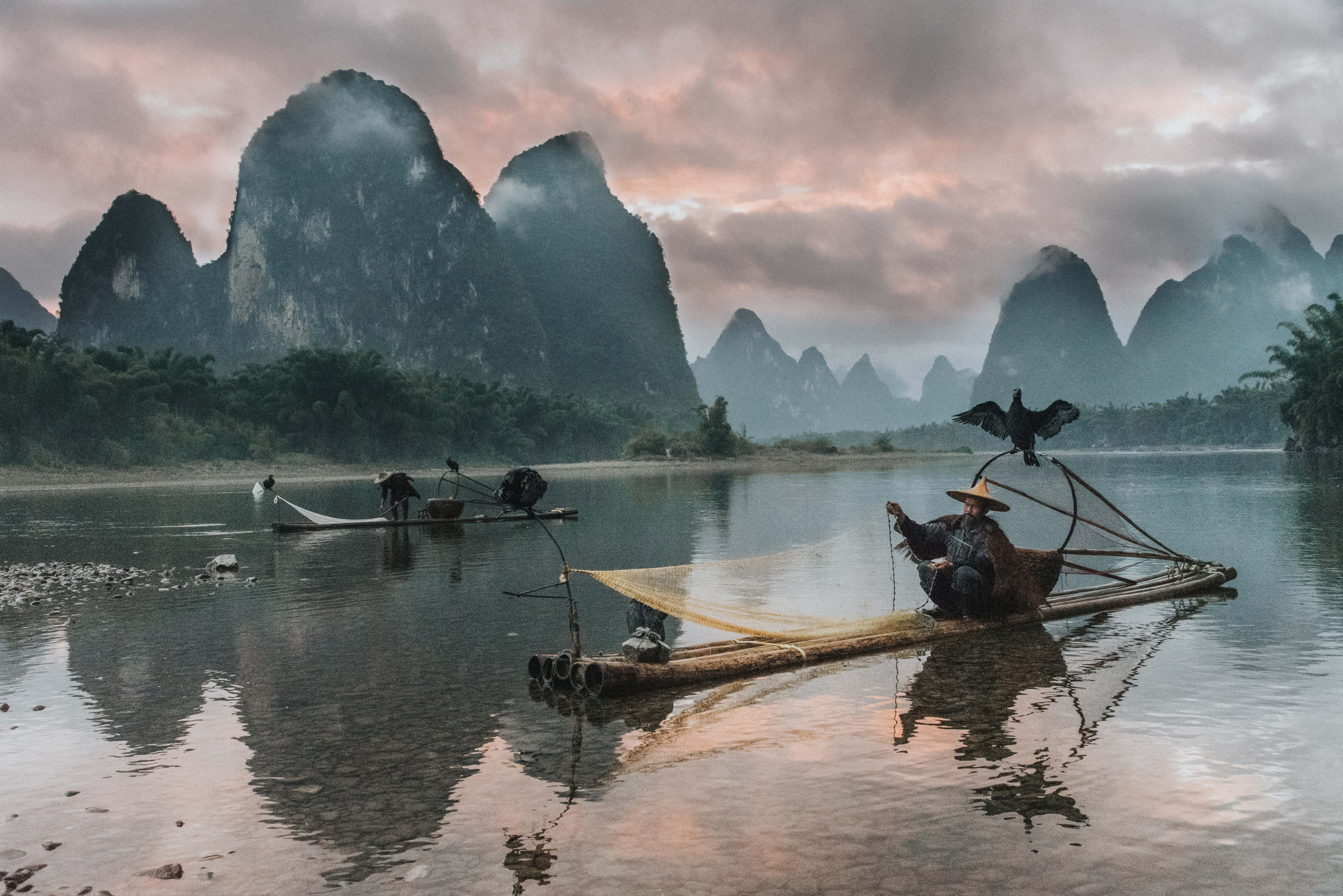 麗江寧靜晨光 壁紙 — 黎明時分，一位漁夫與一隻鸕鶿棲息在竹筏上，霧氣繚繞的喀斯特山脈倒映在平靜的河水之中。
