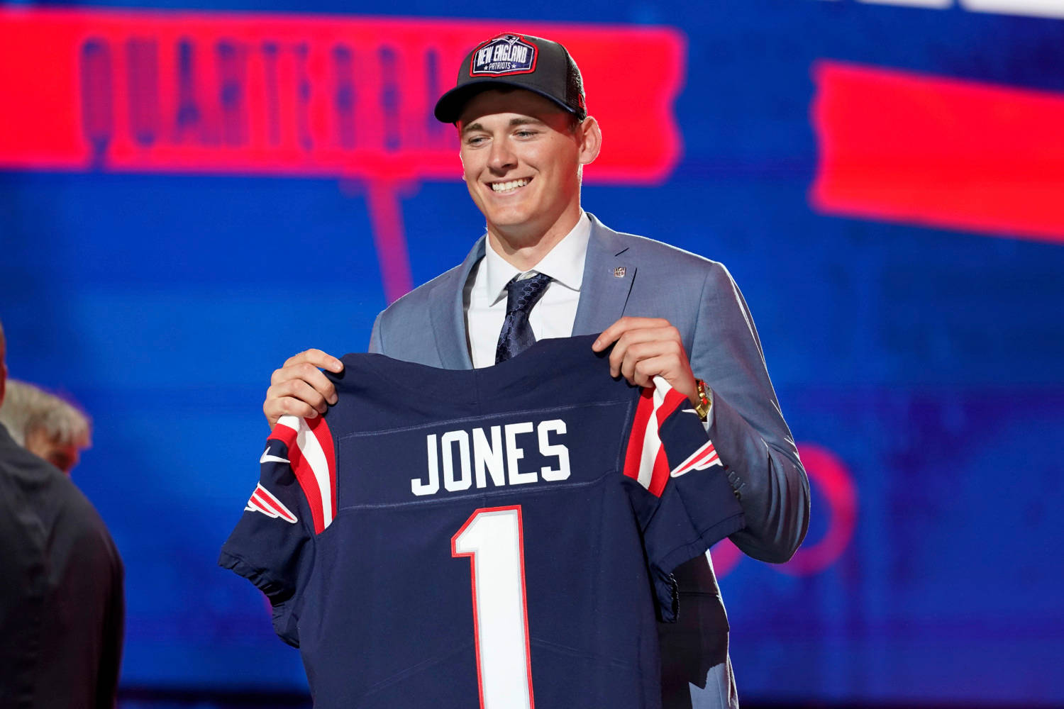 身穿西裝的男子舉起一件印有Jones名字和1號的新英格蘭愛國者隊球衣。