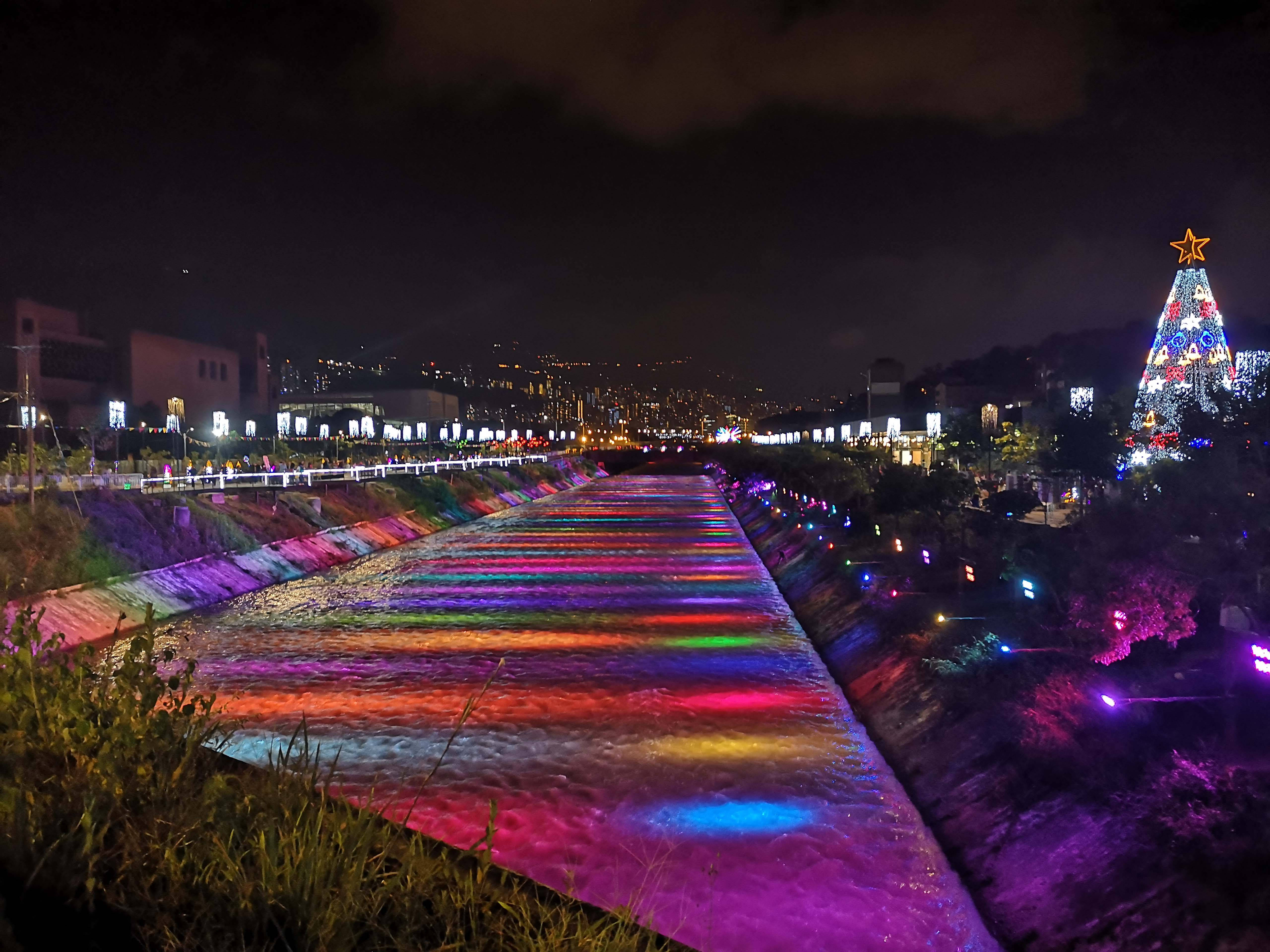 麥德林河畔公園 壁紙 — 一條充滿活力的夜間河流，被彩虹燈光照亮，流經一個節日氣氛濃厚的城市公園，公園內有一棵發光的聖誕樹。