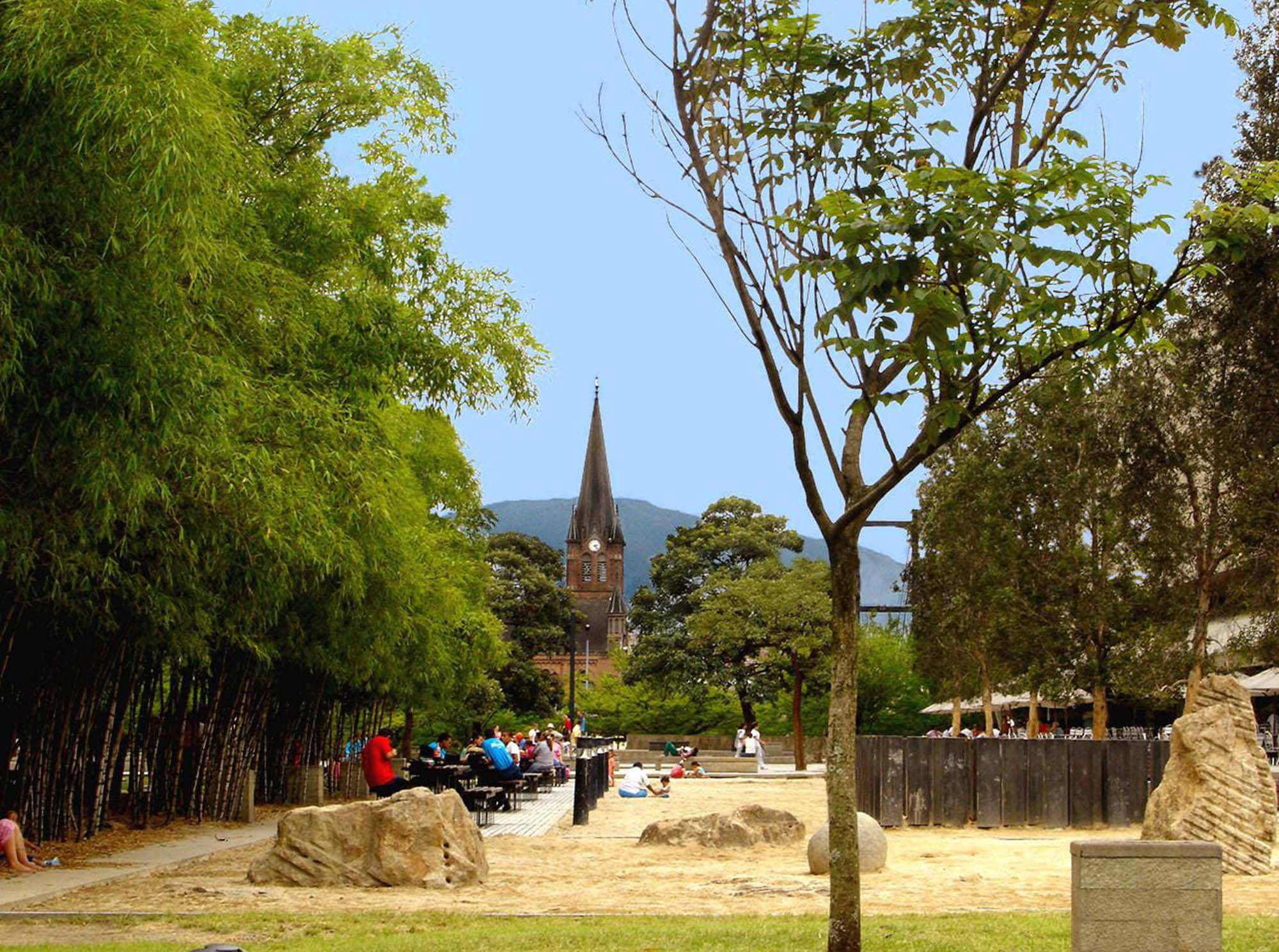 麥德林赤足公園 壁紙 — 陽光明媚的公園場景，人們在竹林附近放鬆，遠處的教堂尖頂映襯著清澈的藍天。