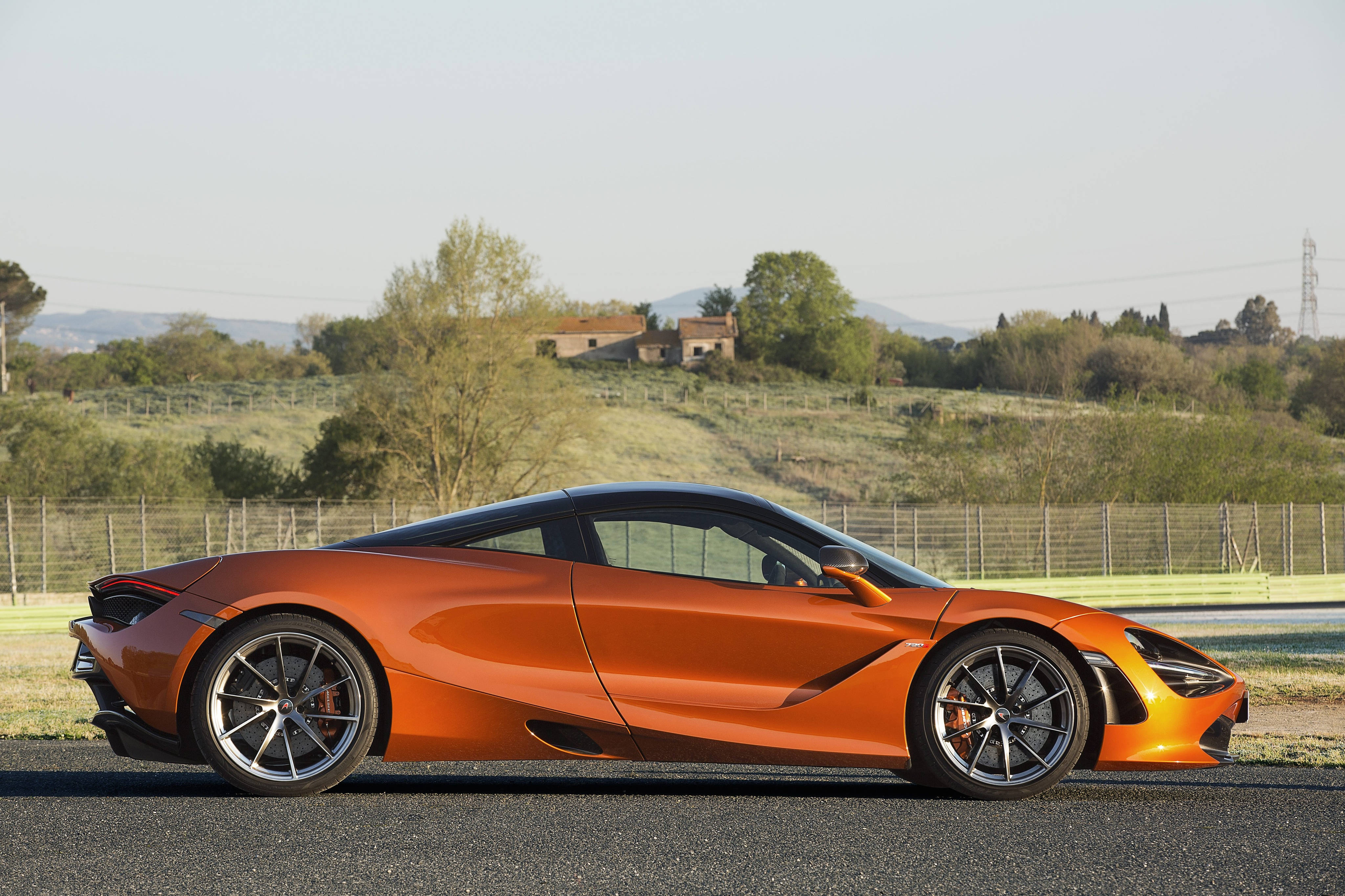 麥拉倫720S 橙色側面 壁紙 — 一輛充滿活力的橙色McLaren跑車在瀝青賽道上側面閃耀，背景是連綿的綠色山丘和遠處的建築物，天空朦朧。
