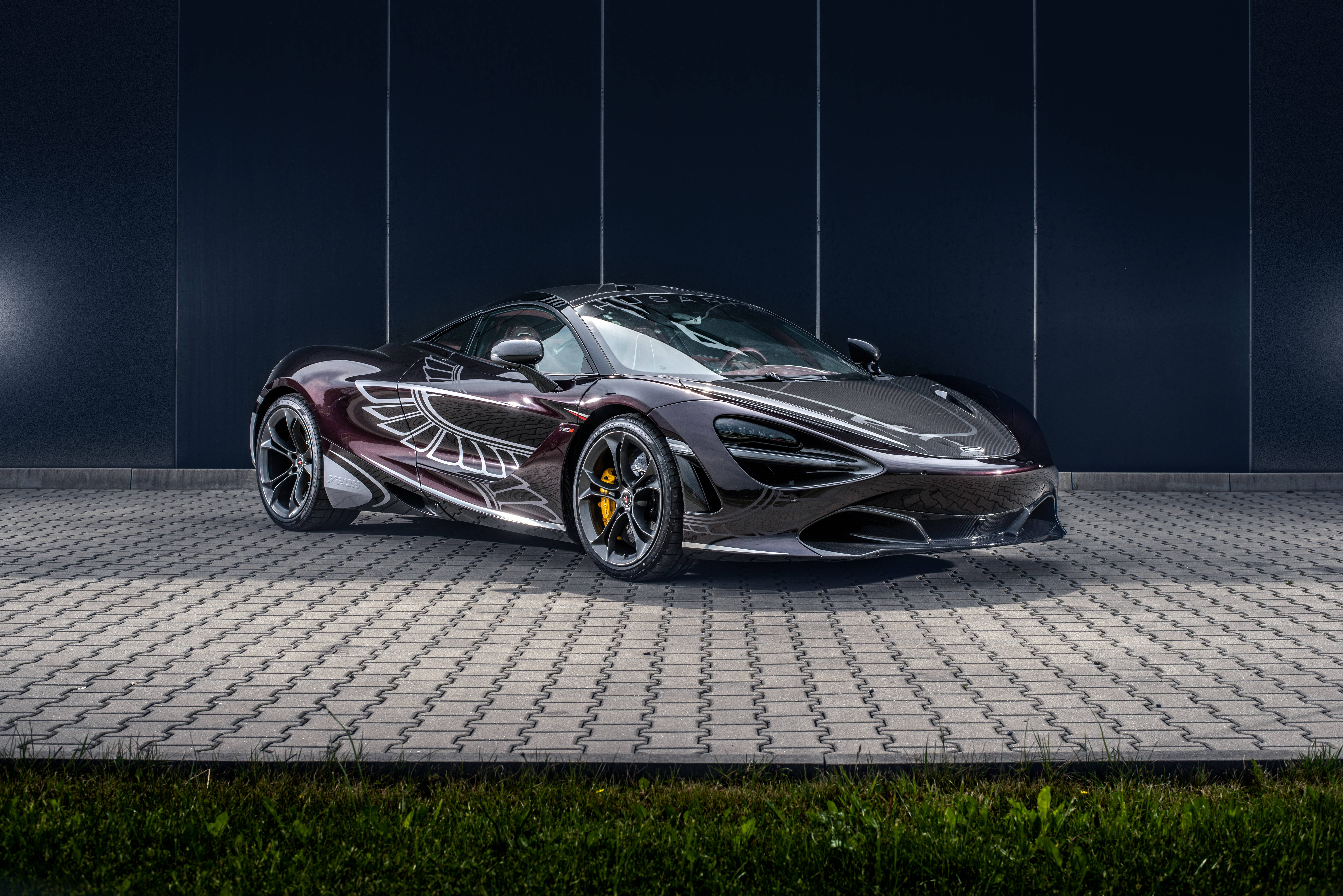 麥拉倫720S 翅膀裝飾 壁紙 — 一架型格黑色McLaren跑車帶有銀色翼形貼紙，停泊在瓷磚鋪設的路面上，背景是深藍色牆壁。