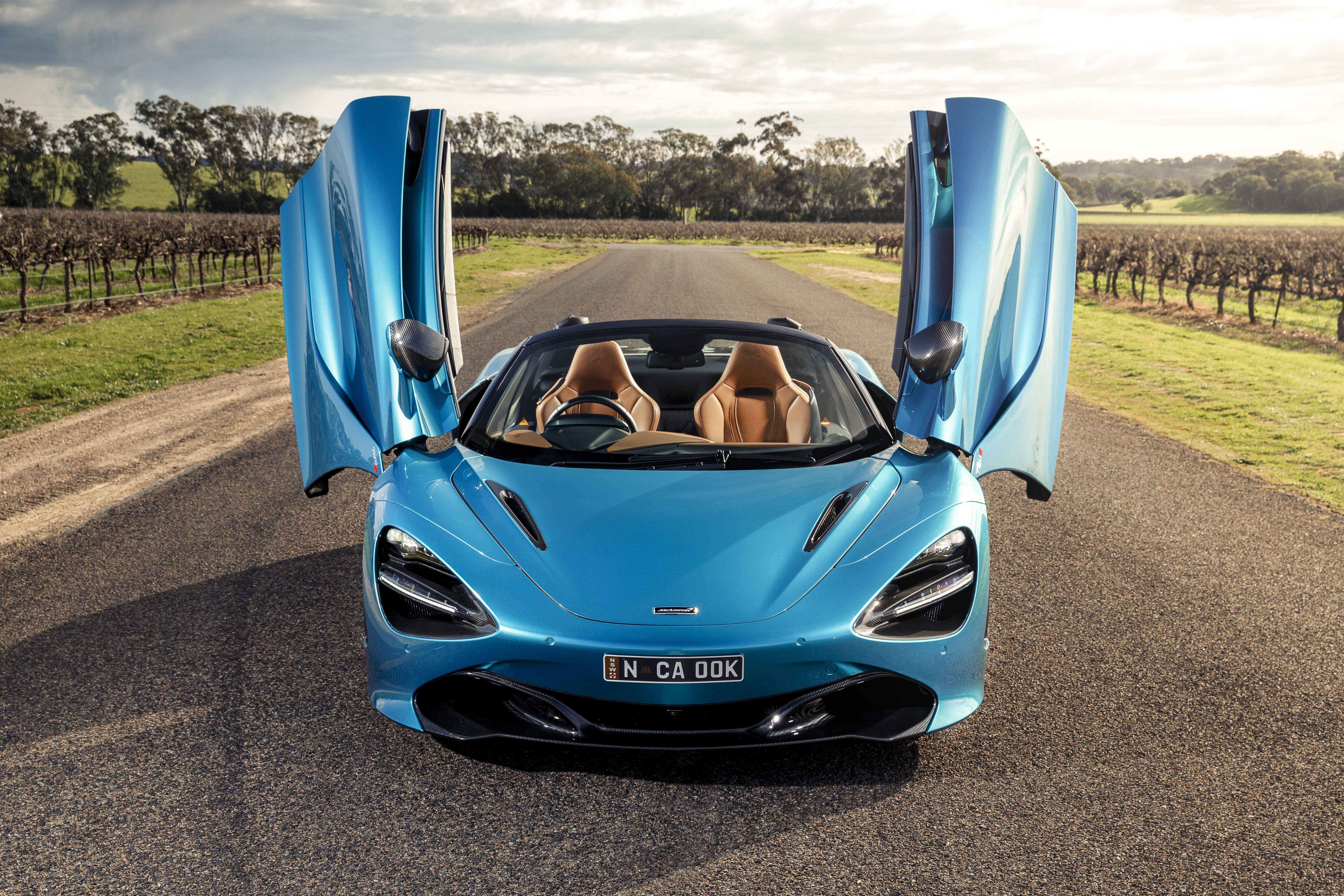 麥拉倫720S 藍色鄉間 壁紙 — 一架鮮藍色McLaren 720S Spider打開鷗翼門，停在一條兩旁種滿葡萄園的鄉村道路上，天空柔和。