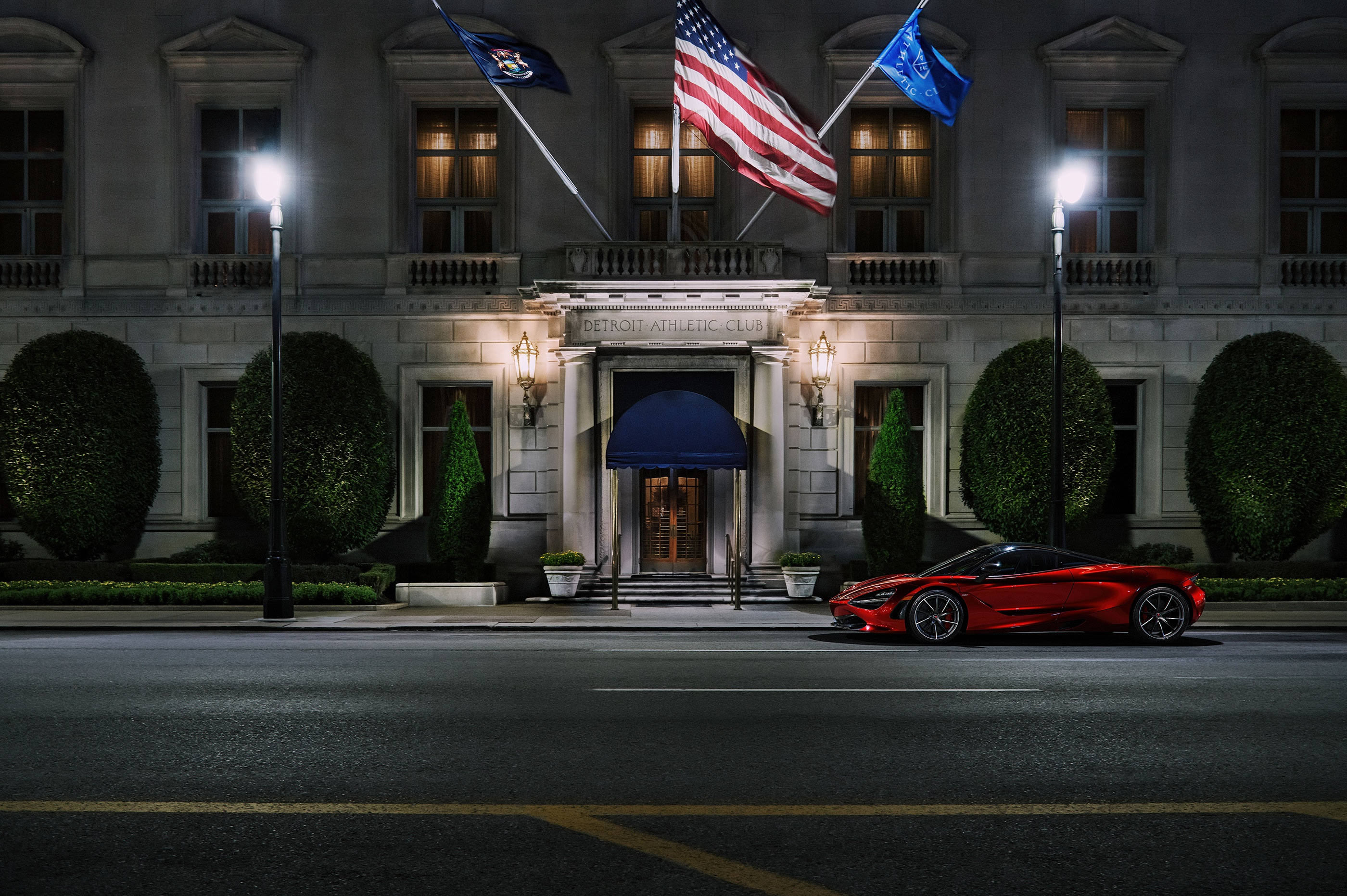 麥拉倫720S 運動俱樂部 壁紙 — 一輛時尚的紅色跑車在夜間停泊在一座宏偉的燈光建築前，建築物有旗幟和修剪整齊的樹籬。