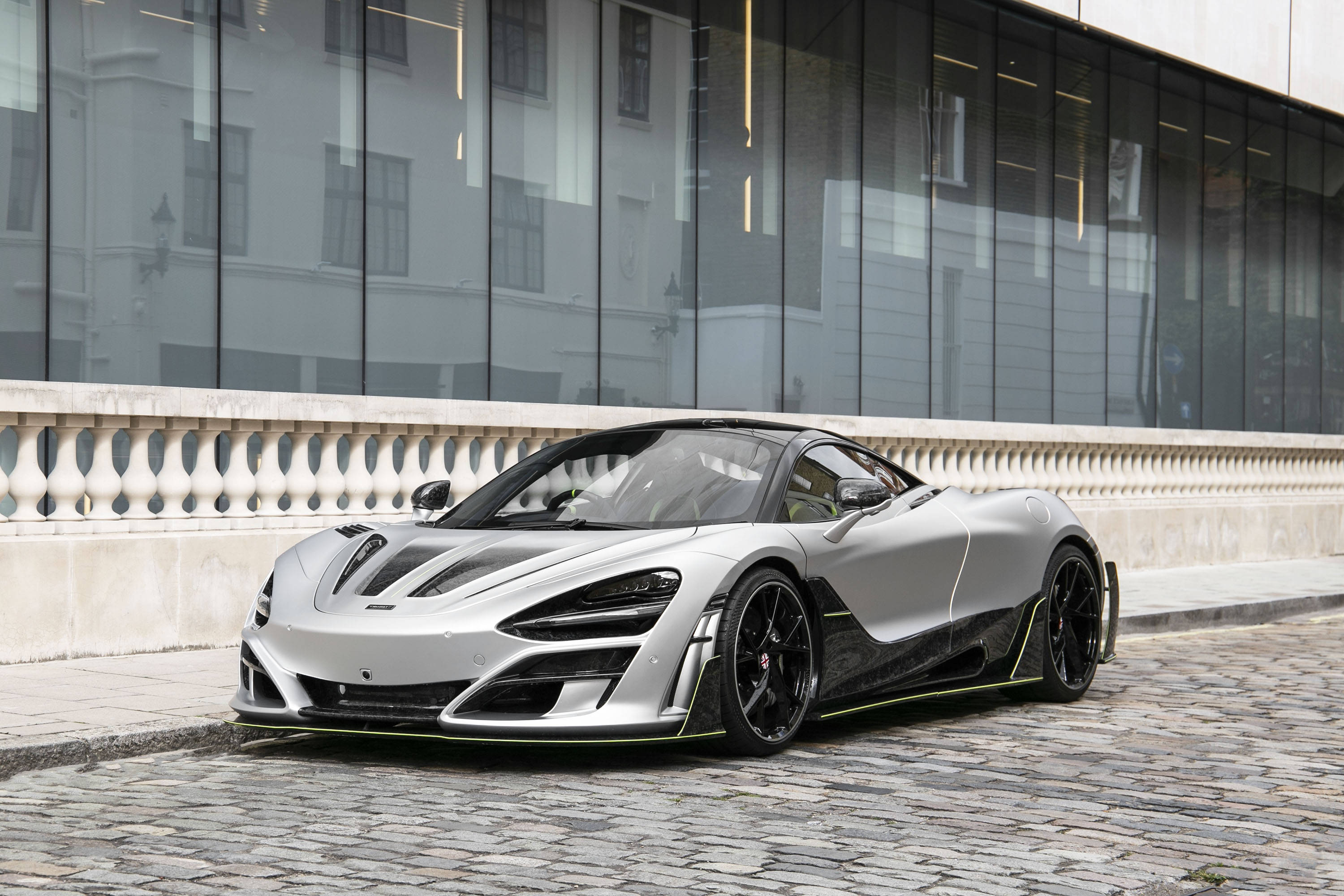 麥拉倫720S 銀色 壁紙 — 一輛時尚的銀色麥拿侖跑車，帶有黑色飾邊，停泊在現代玻璃建築旁的鵝卵石街道上。