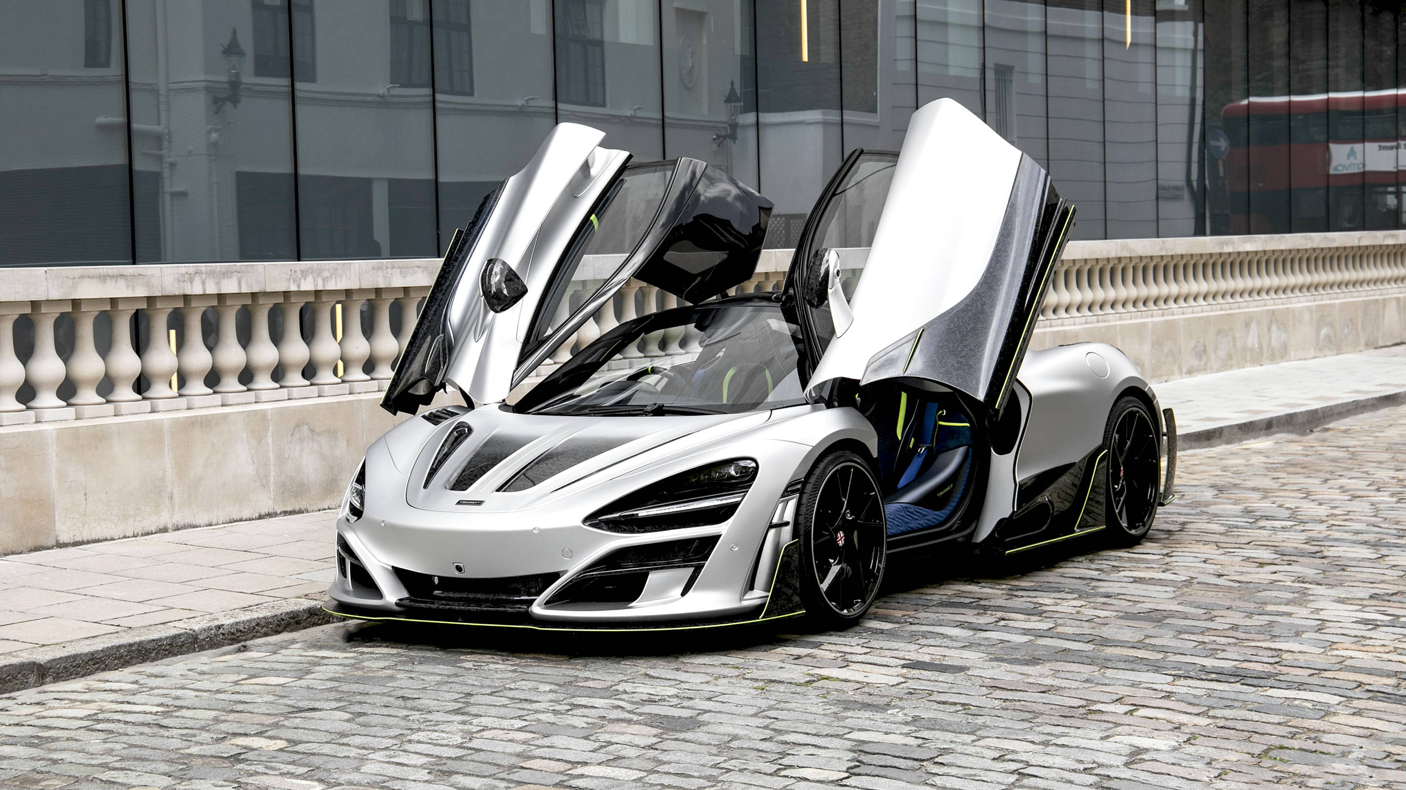 麥拉倫720S 銀色城市 壁紙 — 一架銀色麥拿倫Senna，鷗翼車門打開，停在鵝卵石街道上，旁邊是現代玻璃建築。
