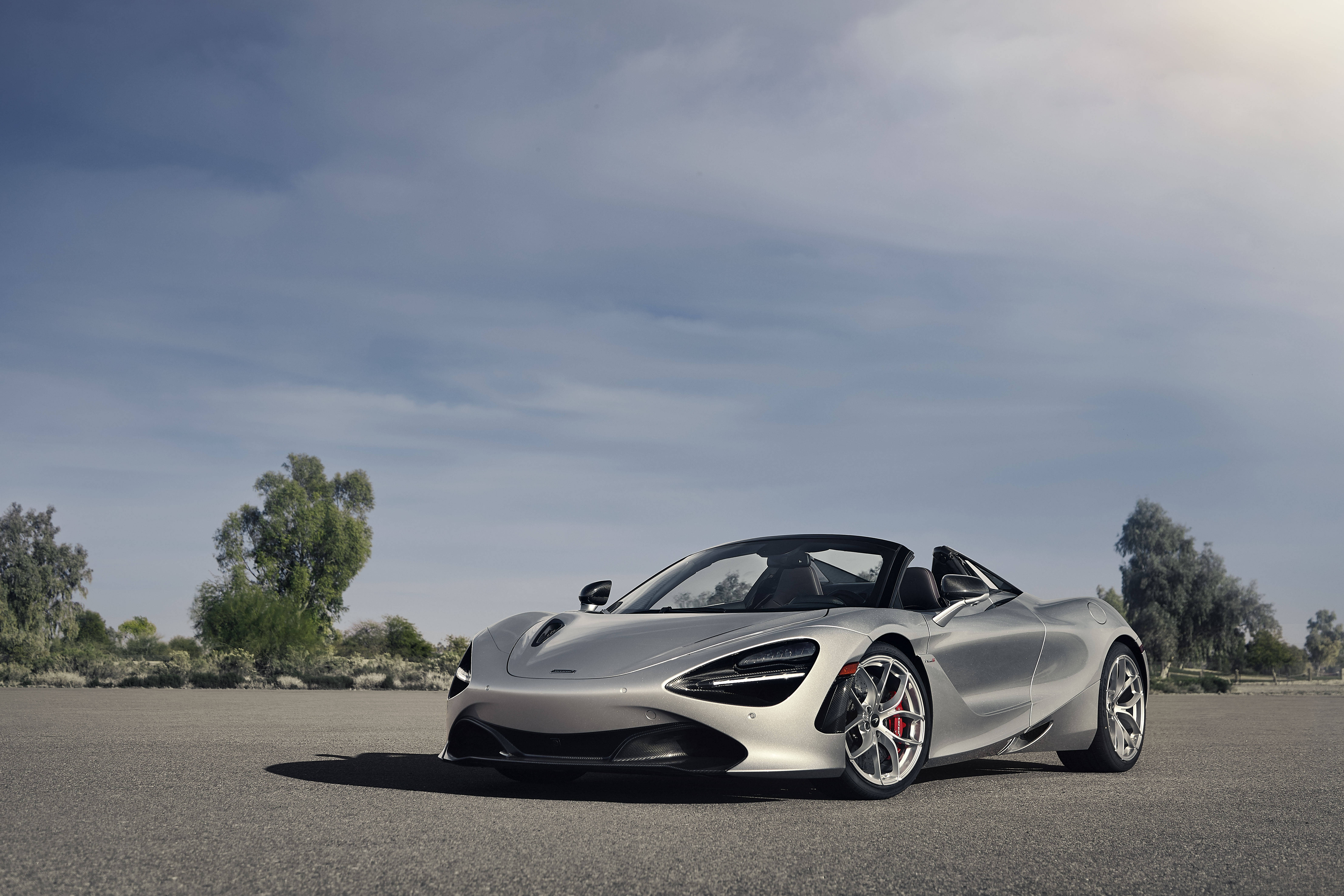 麥拉倫720S 銀色戶外 壁紙 — 一架時尚的銀色McLaren 720S Spider開篷跑車停泊在開闊的瀝青路上，天空部分多雲，背景有稀疏的樹木。
