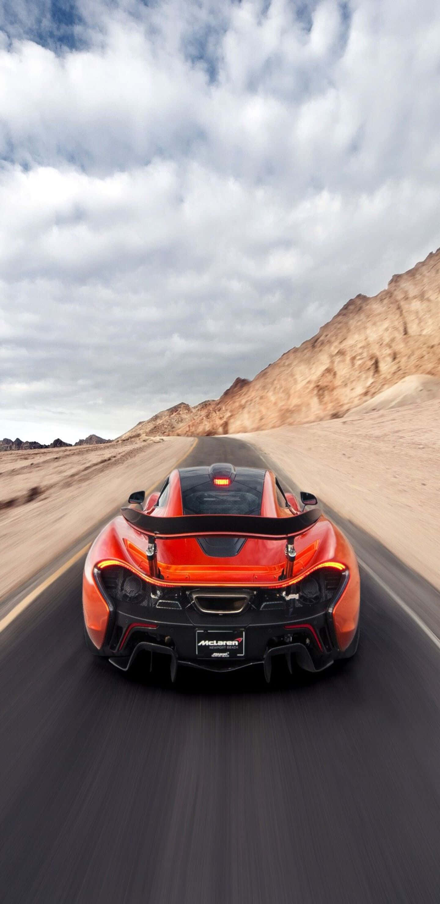 麥拉倫720S天空背景（Pixel 3XL格式） 壁紙 — 一輛火橙色的McLaren跑車在雲層密布的天空下，沿著一條有岩石山丘的沙漠公路飛馳。