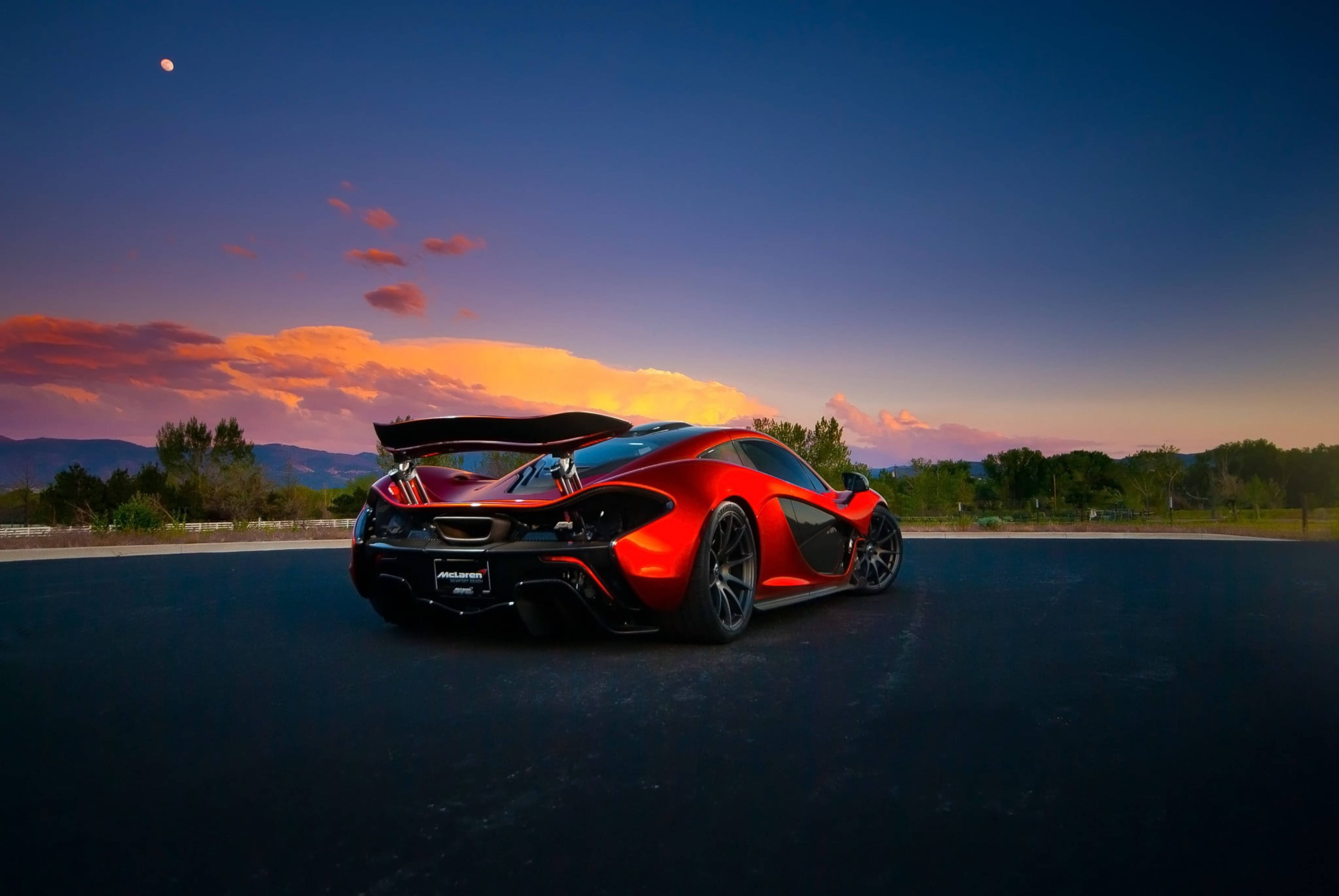 麥拉倫P1跑車後視圖 壁紙 — 一架鮮紅色的McLaren跑車，尾翼升起，停在日落時分的深色瀝青路上，背景是遠山和色彩繽紛的天空，還有一輪可見的月亮。