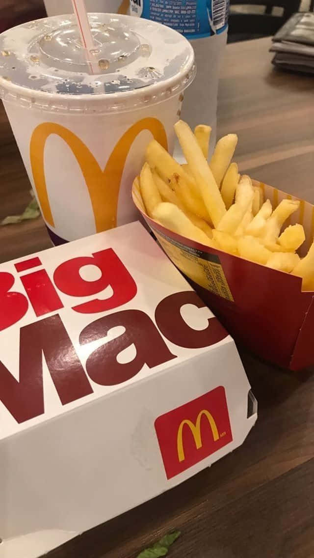 麥當勞巨無霸配薯條餐點圖片 壁紙 — 木桌上麥當勞巨無霸盒和薯條旁邊放著飲料杯的特寫。