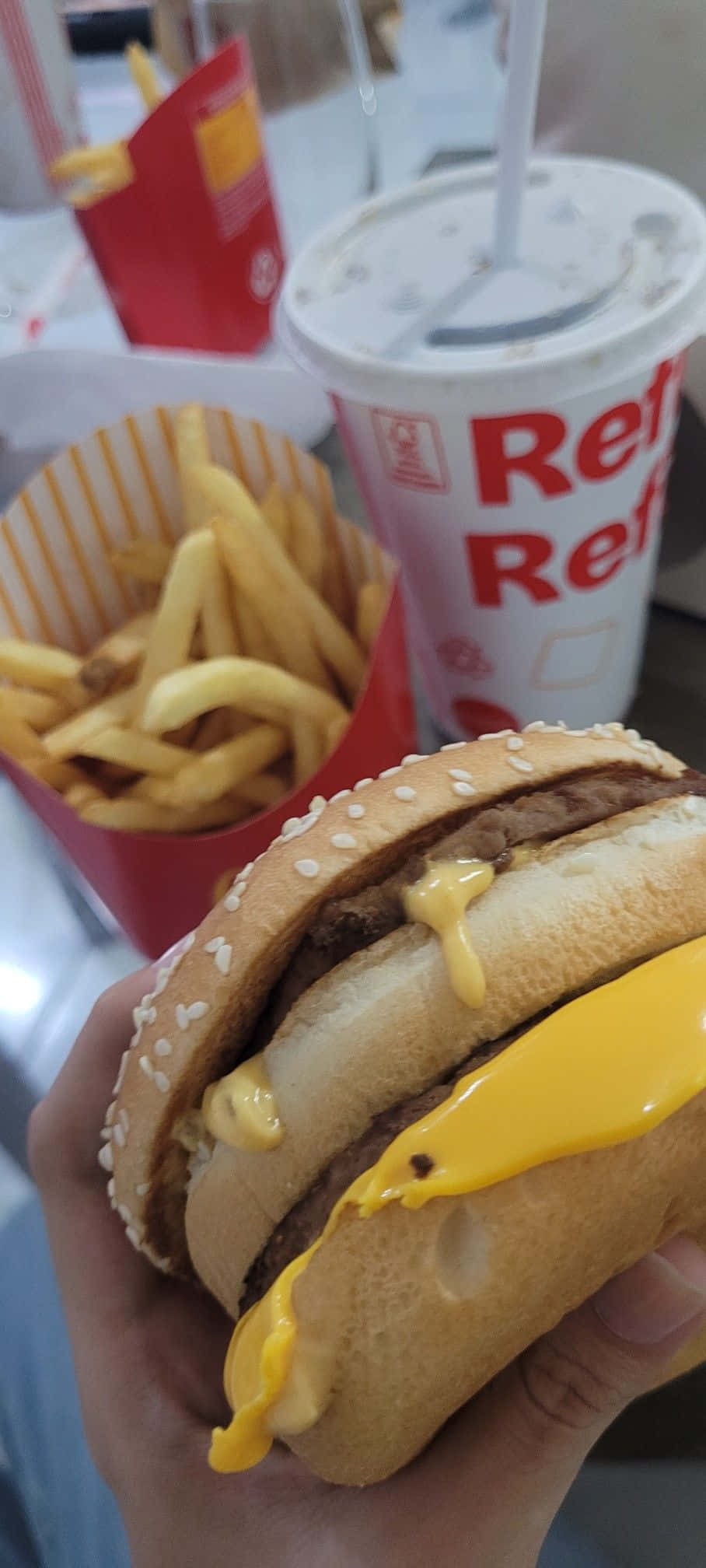 麥當勞漢堡與薯條美食圖 壁紙 — 一隻手拿著滴汁的芝士漢堡，配薯條和飲品，身處快餐環境中。