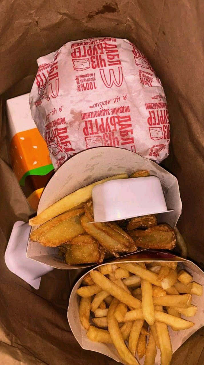 麥當勞薯條與漢堡袋圖 壁紙 — 快餐餐點的近距離特寫，包括一個用品牌紙包裹的漢堡和兩份放在紙籃裡的金黃色薯條。