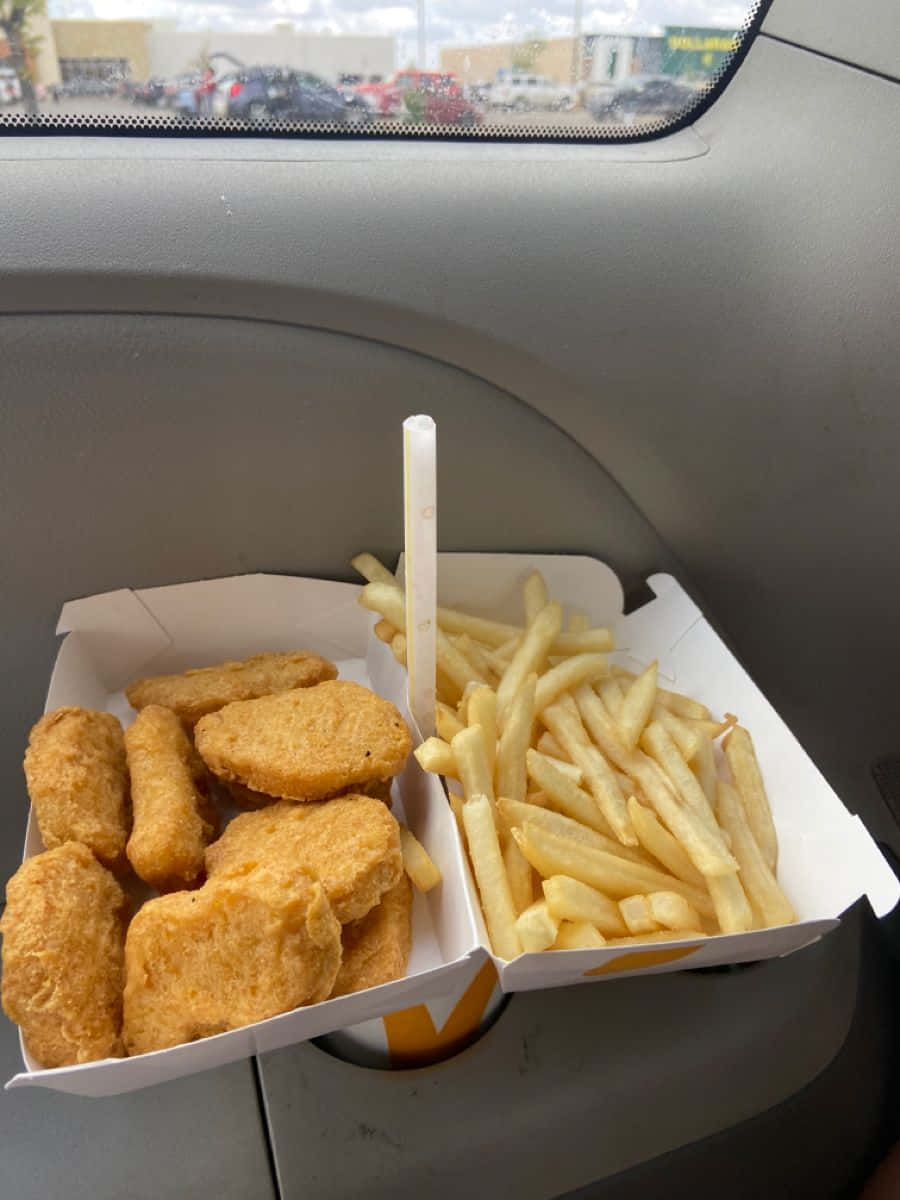 麥當勞雞塊與薯條餐點圖片 壁紙 — 金黃色的炸雞塊和薯條放在白色盒子裡，擱在汽車杯架上，可看到停車場景色。