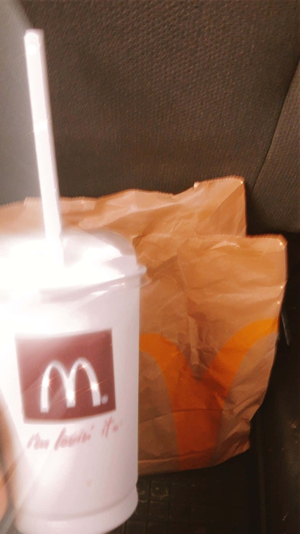 麥當勞飲品的美感呈現 壁紙 — 一個麥當勞飲料杯連飲管放在車座上，旁邊有一個皺巴巴的紙袋。