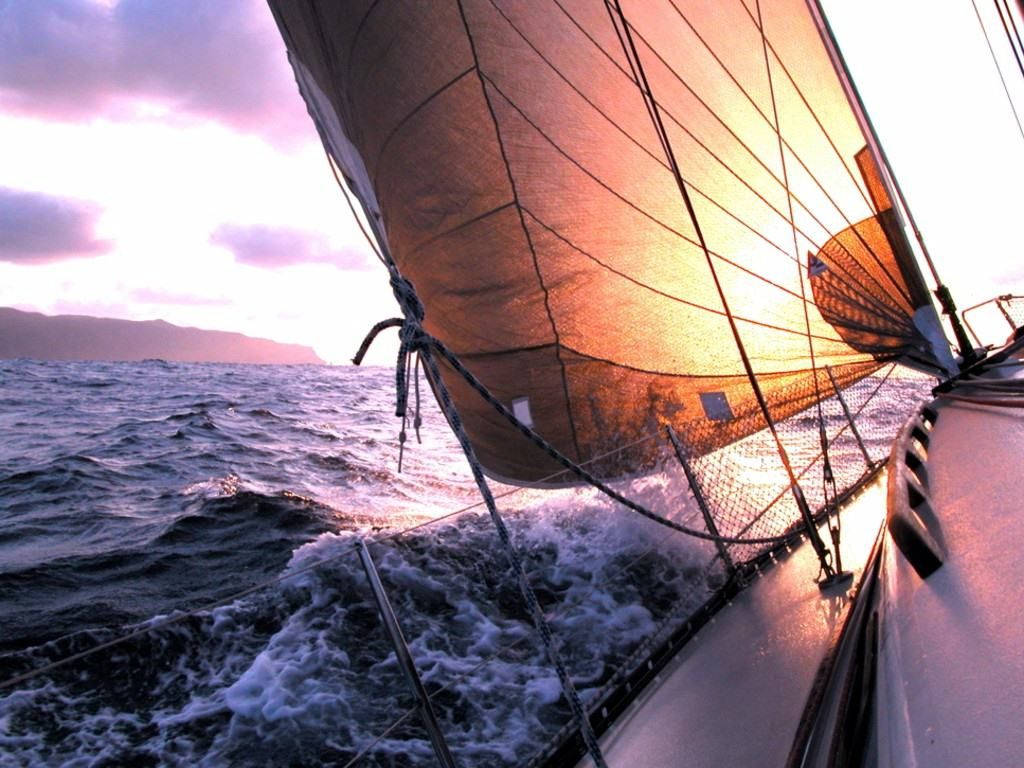 黃帆駛過夕照 壁紙 — 一艘帆船在日落時分穿過波濤洶湧的海浪，帆在溫暖的光線中閃耀，映襯著紫色的天空。