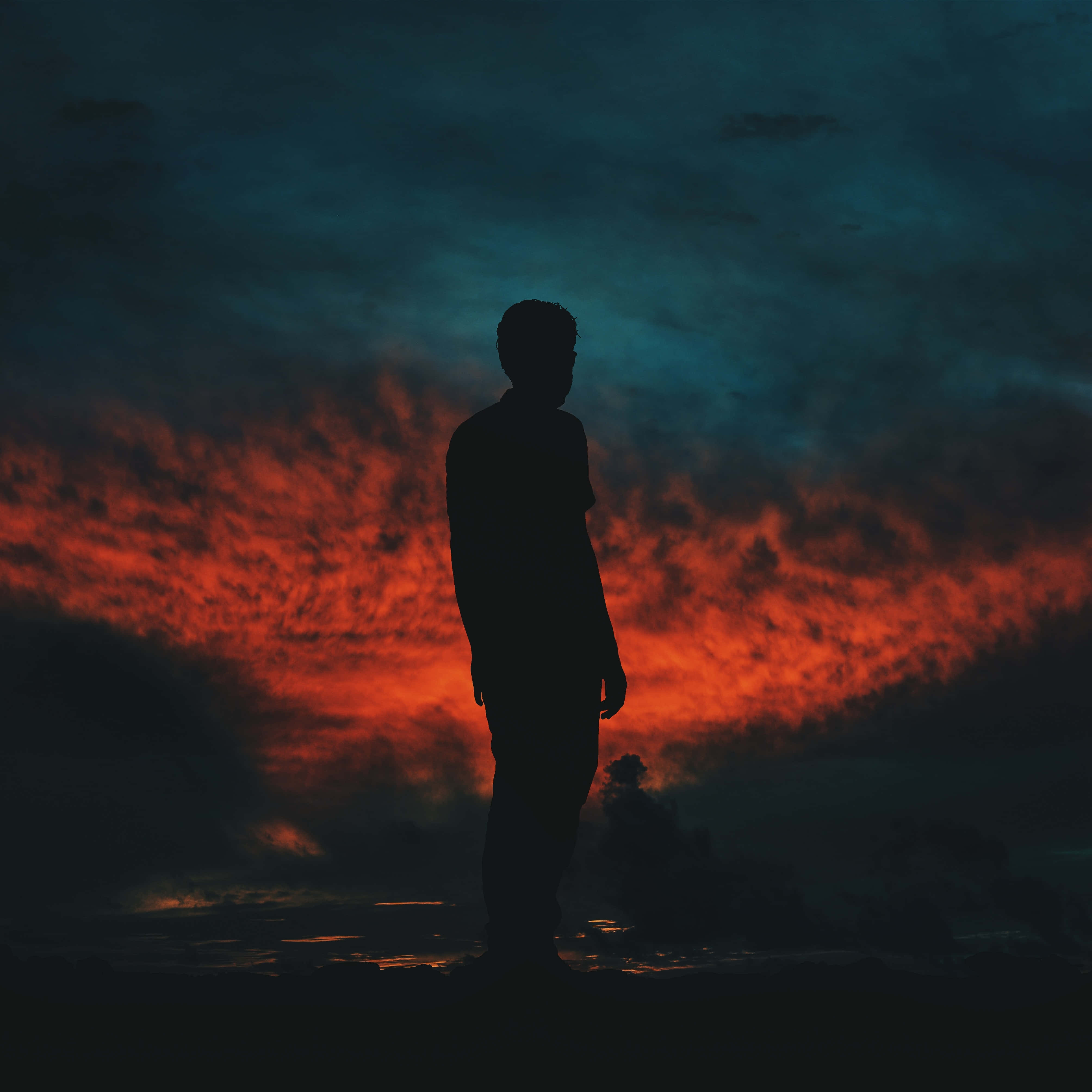 Silhouette 桌布 壁紙 — 黃昏時分，一個孤獨的人影站在一片火橙色和深青色雲彩交織的戲劇性天空下。