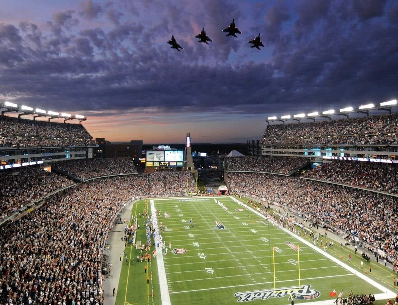 Football Stadium 桌布 壁紙 — 黃昏時分，一個擠滿人的足球場上空，四架戰鬥機以編隊飛行，背景是紫色的天空。