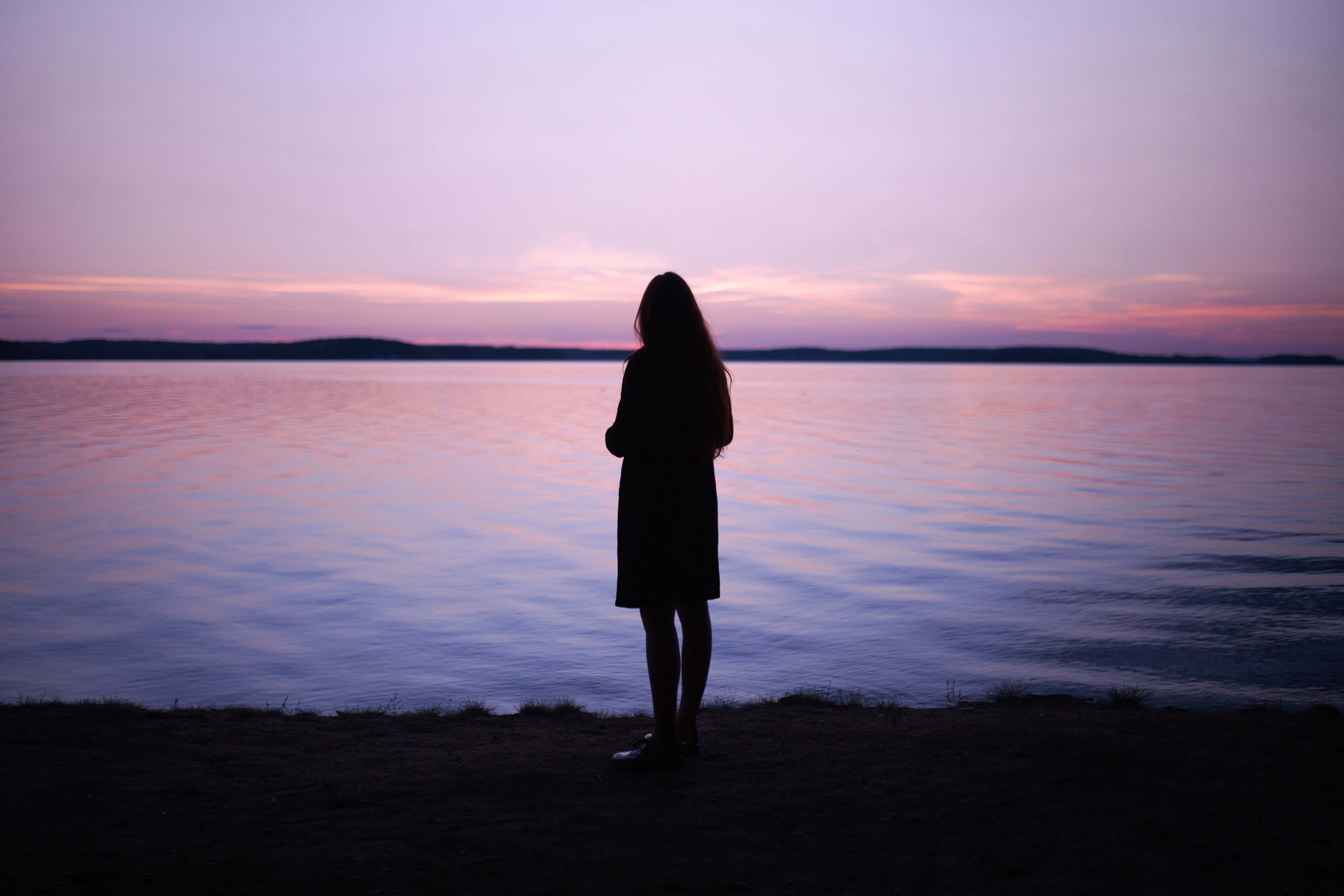 Sunset Silhouette 桌布 壁紙 — 黃昏時分，一名孤獨女子站在平靜的水邊，天空呈現柔和的紫色和粉紅色剪影。