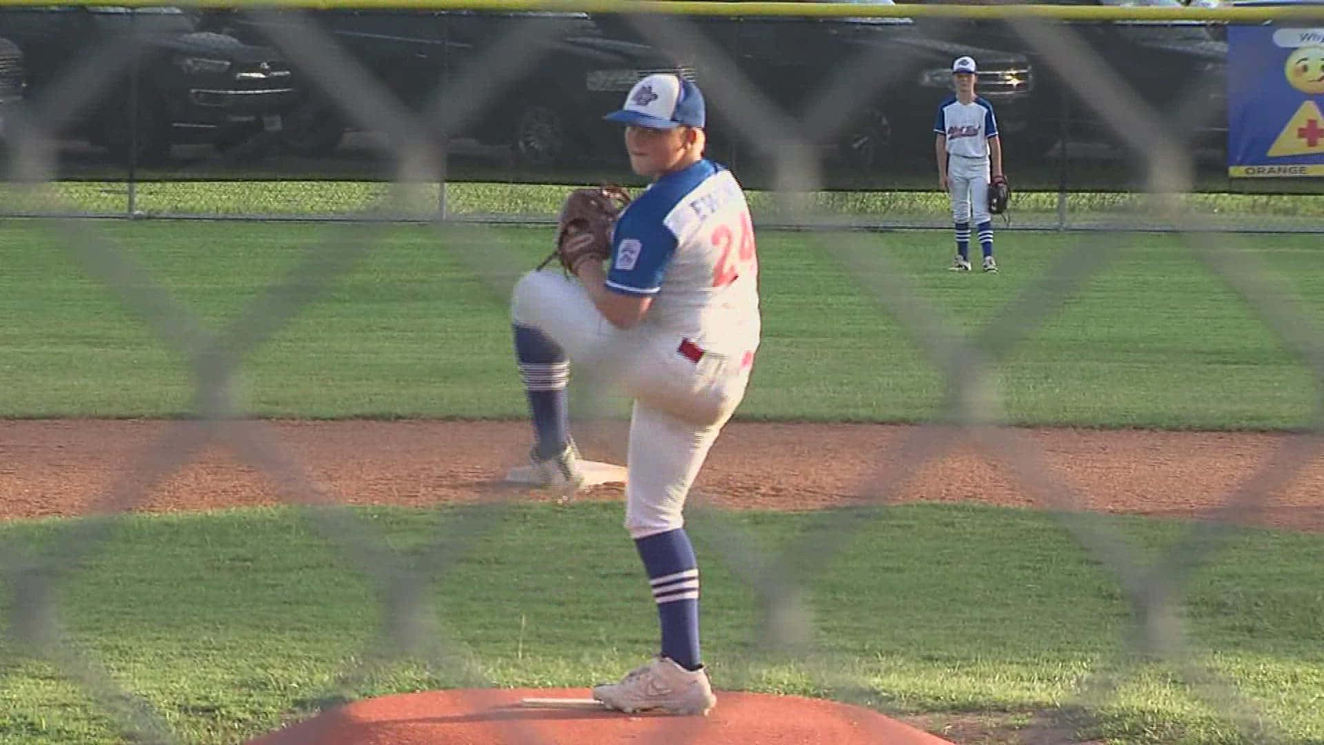 Little League Baseball 桌布 壁紙 — 黃昏時分，一名身穿藍白球衣的年輕棒球投手透過鐵絲網在投手丘上揮臂準備投球。