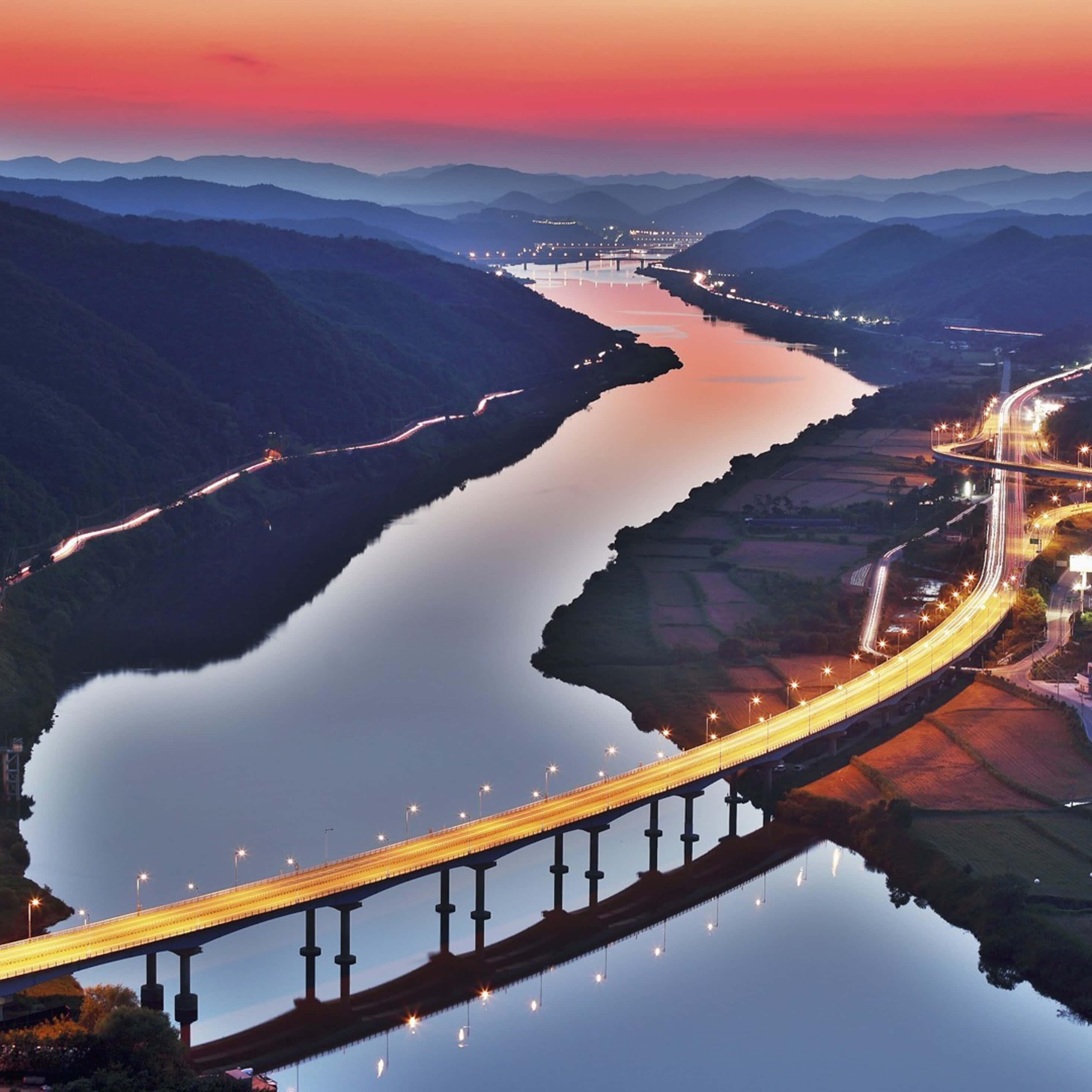 South Korea 桌布 壁紙 — 黃昏時分，一條發光的高速公路橋樑橫跨蜿蜒的河流，層層山脈在火紅的粉紅色和橙色天空下的鳥瞰圖。