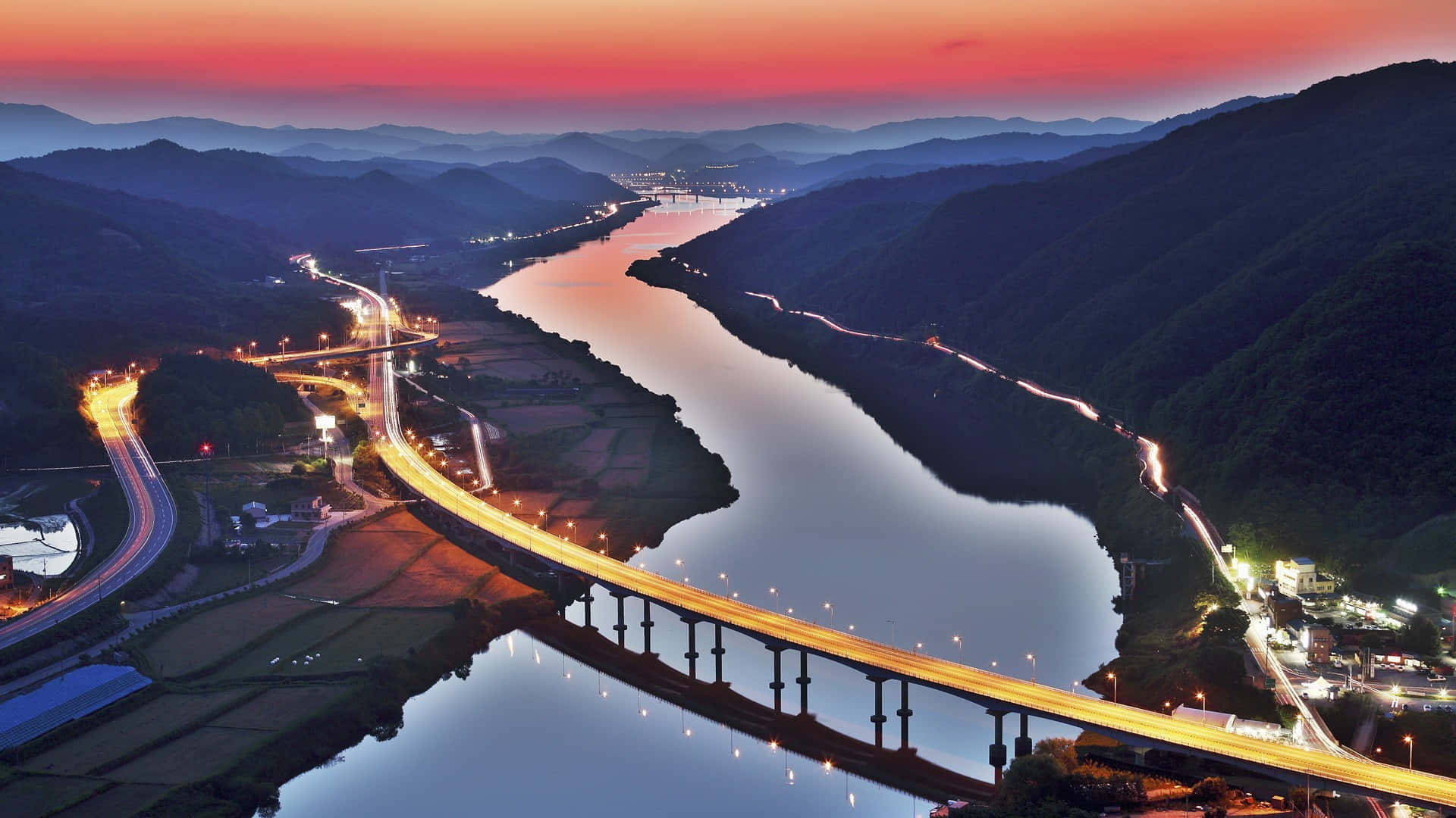 South Korea 桌布 壁紙 — 黃昏時分，一條發光的高速公路橋樑橫跨蜿蜒的河流，層層疊疊的山脈在火紅的粉紅色和橙色天空下的鳥瞰圖。