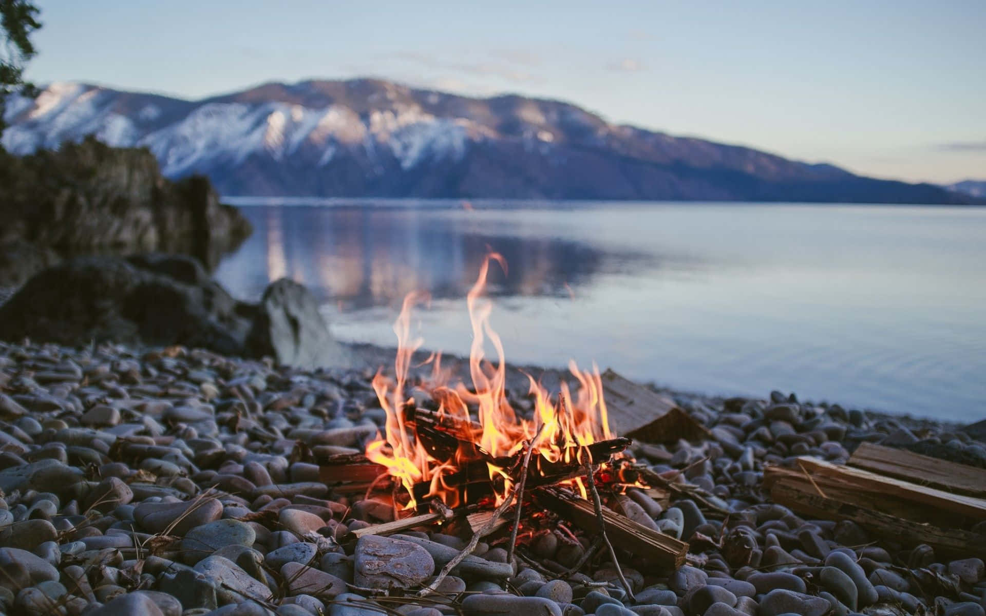 Campfire 桌布 壁紙 — 黃昏時分，卵石海灘上噼啪作響的營火熊熊燃燒，背景是白雪覆蓋的山脈和寧靜的湖泊。