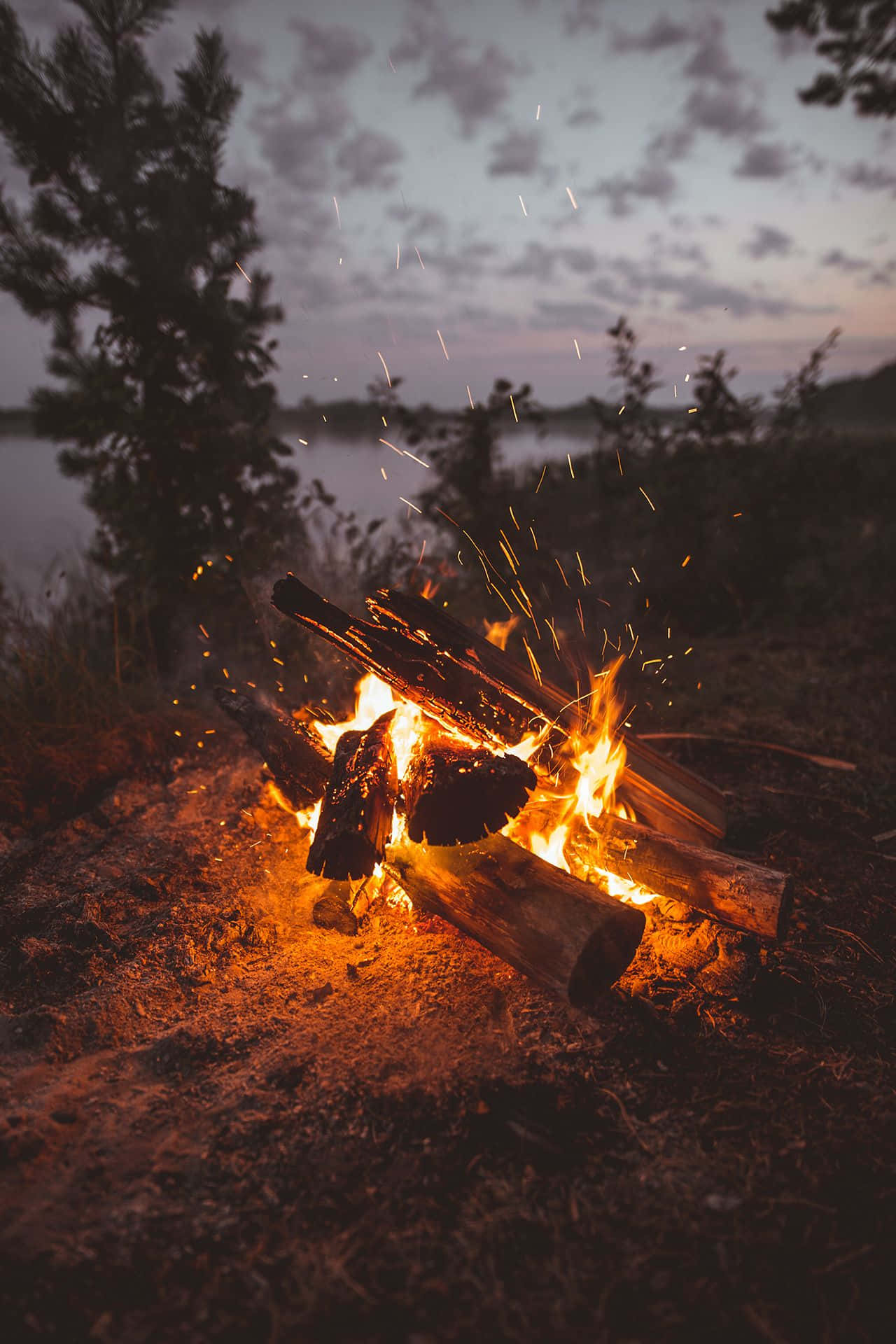 Cozy Fire 桌布 壁紙 — 黃昏時分，噼啪作響的營火將金色火花送入寧靜湖邊的暮色天空。