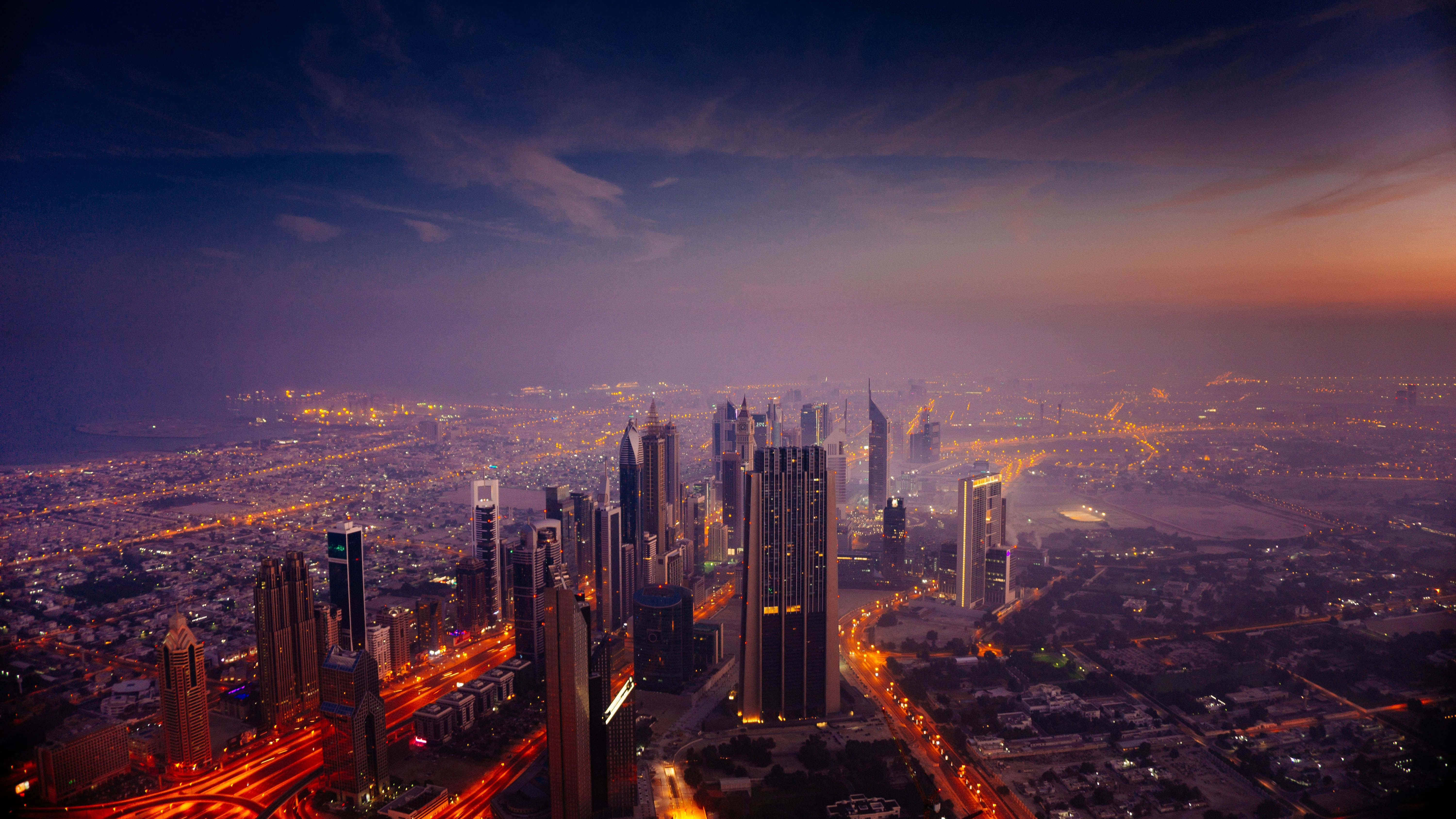 Dubai 桌布 壁紙 — 黃昏時分，廣闊城市景觀的鳥瞰圖，摩天大樓燈火通明，道路在紫橙色天空下閃閃發光。