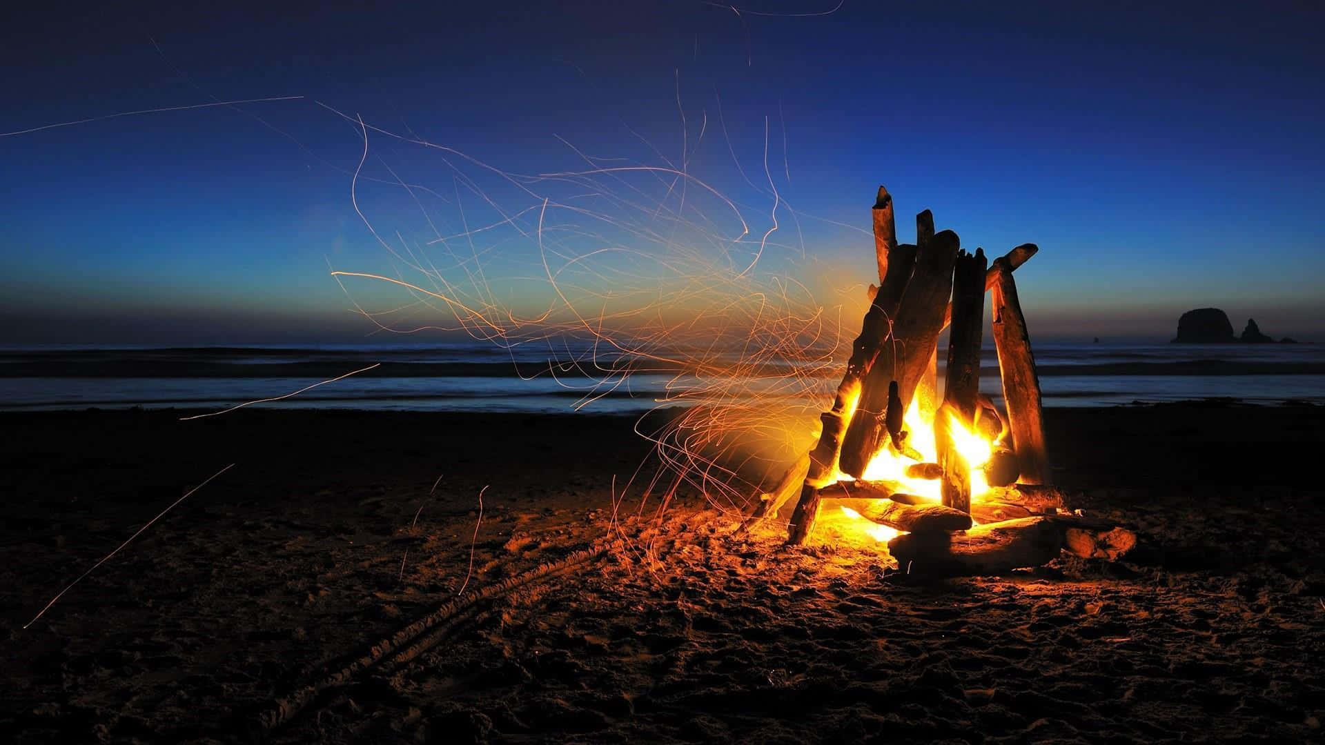 Beach Bonfire 桌布 壁紙 — 黃昏時分，沙灘上熊熊燃燒的營火，發光的火花在深藍色的天空和遠處的海洋中飛舞。